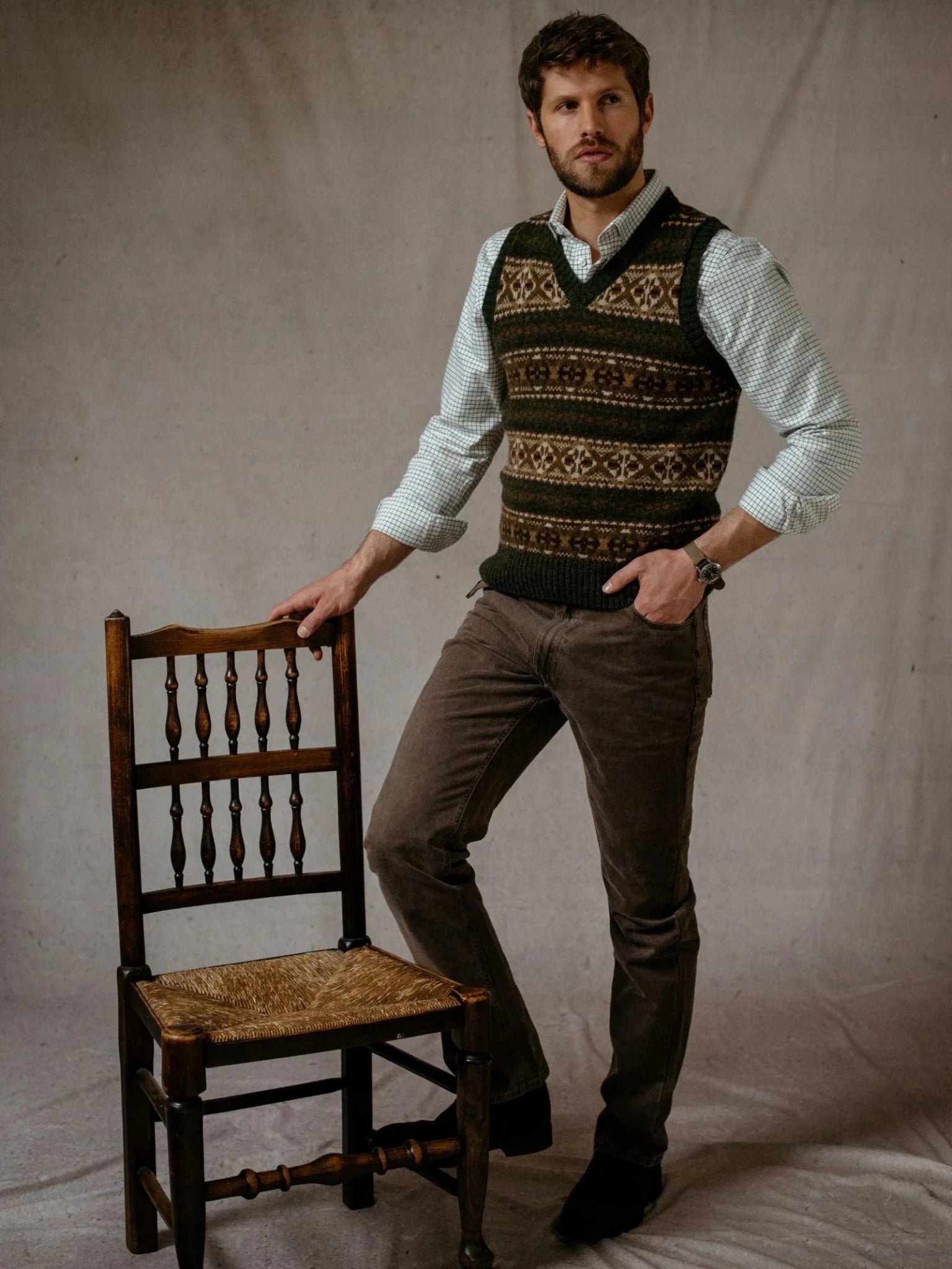 A man wearing the Campbell’s of Beauly Fairisle Slipover over a dress shirt and brown pants stands with one hand on a wooden chair against a plain backdrop.
