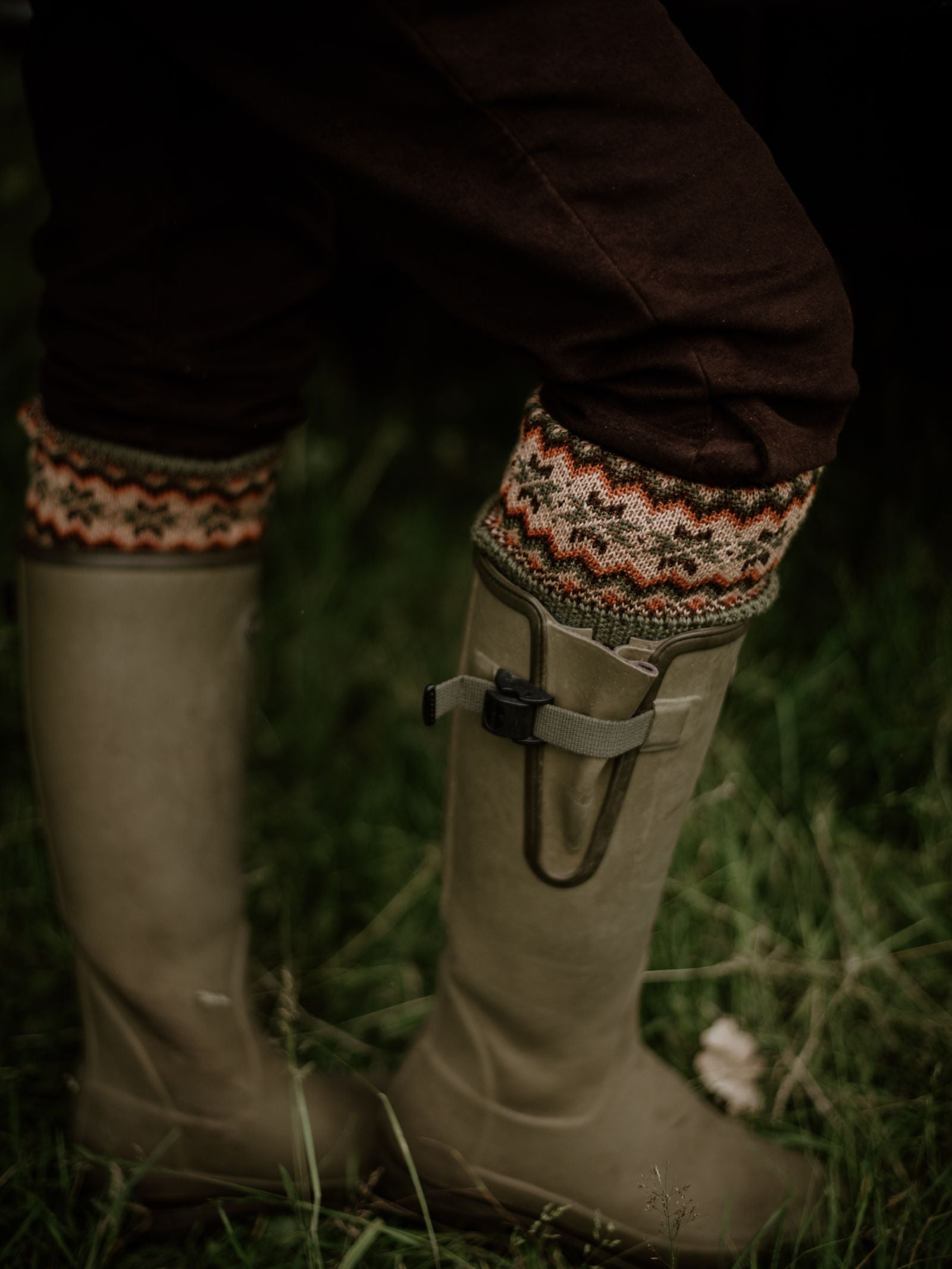 A person stands on grass wearing Campbells of Beauly Moleskin Plus Twos, woolen leg warmers, and olive green buckle-fastened rubber boots; the classic knit pattern completes this timeless countryside look.