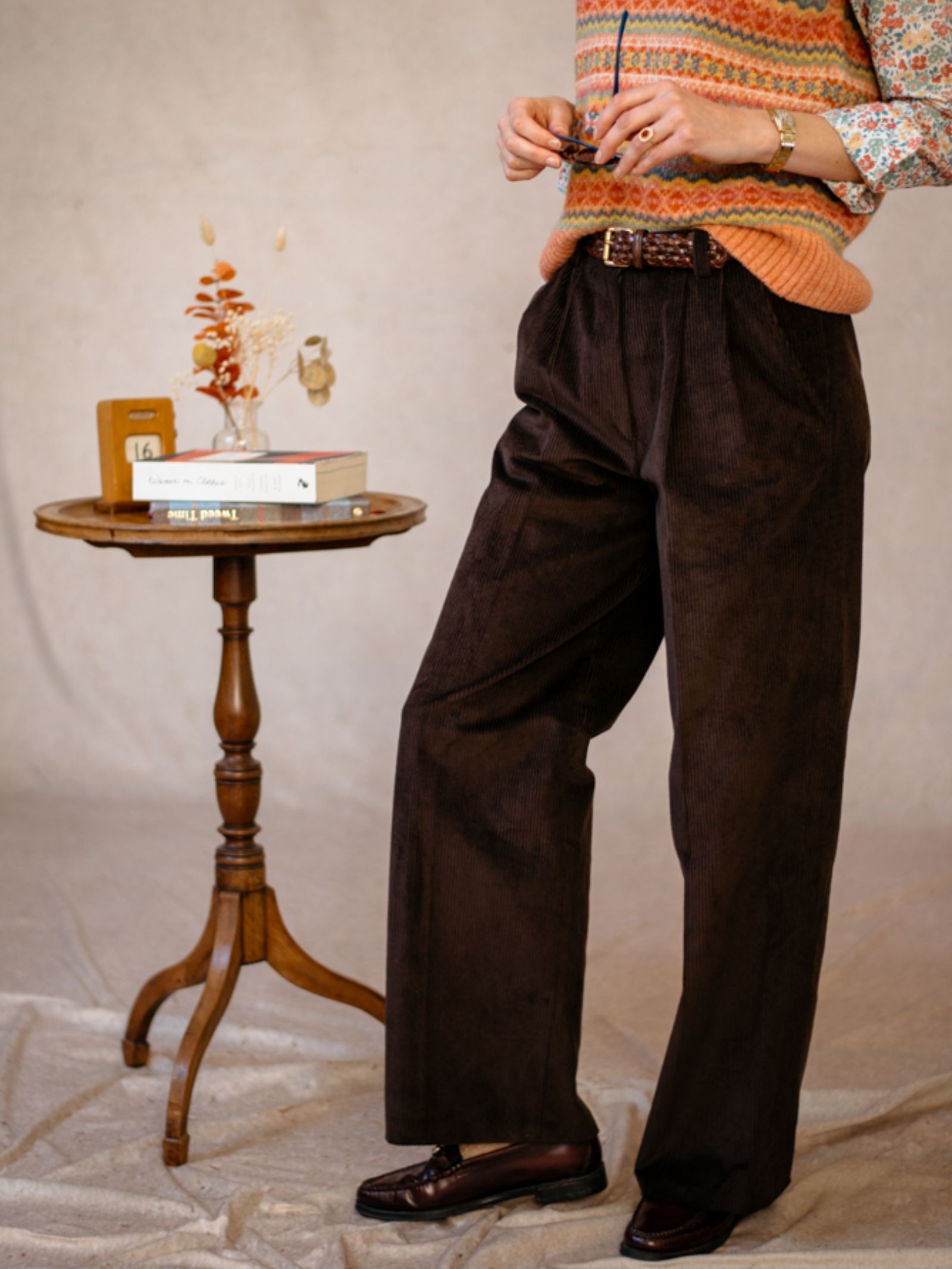 Wearing Campbell's of Beauly Auld Stock Wide Leg Cords, a person stands by a small round wooden table topped with dried flowers, books, and eyeglasses. They pair the cords with a patterned sweater vest, floral shirt, and brown loafers. Face not shown.