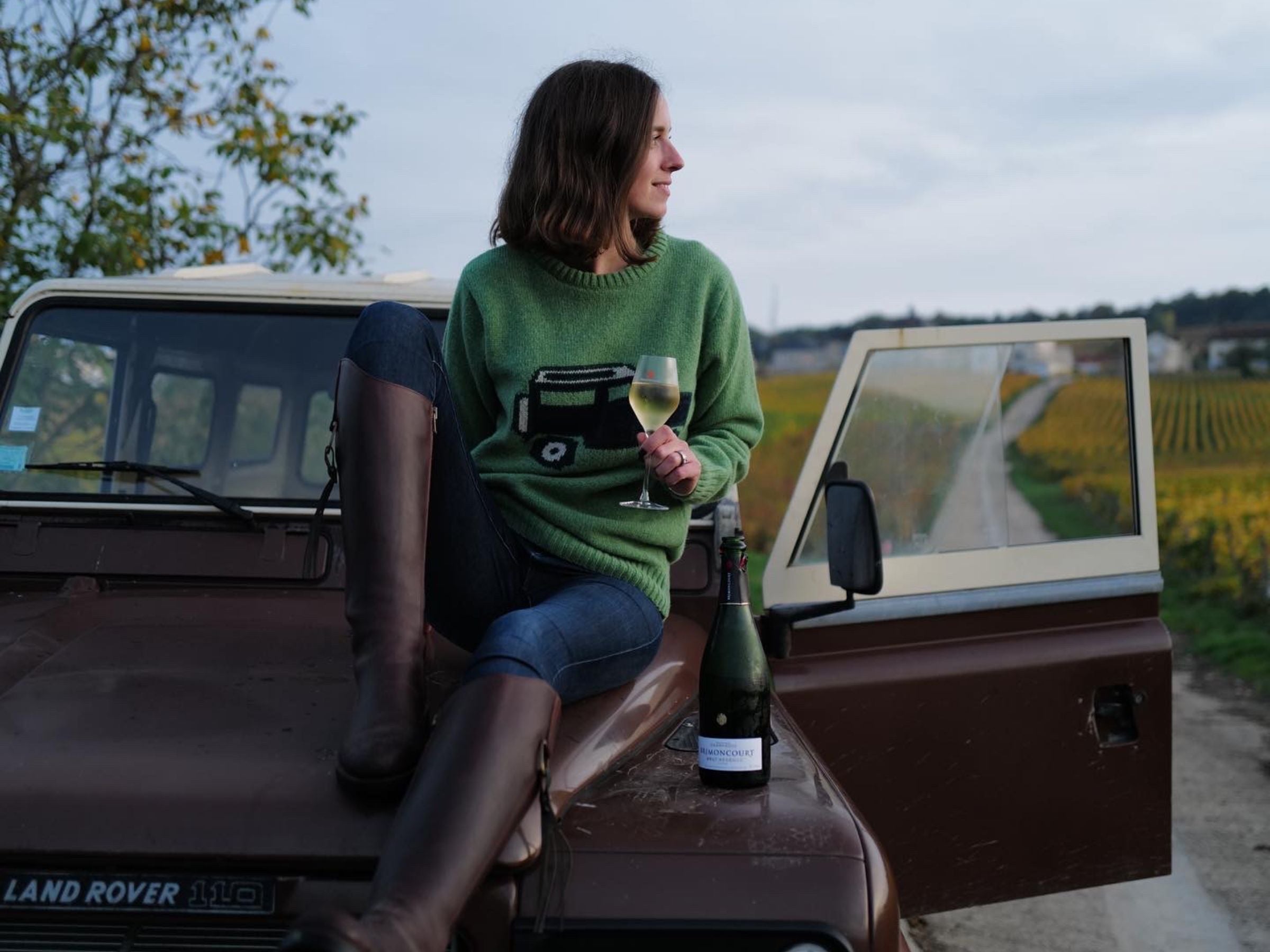 A woman in a green sweater and brown boots sits on a brown Land Rover with an open door, holding a glass of white wine. A wine bottle rests nearby. In the background, there are fields and a country road.