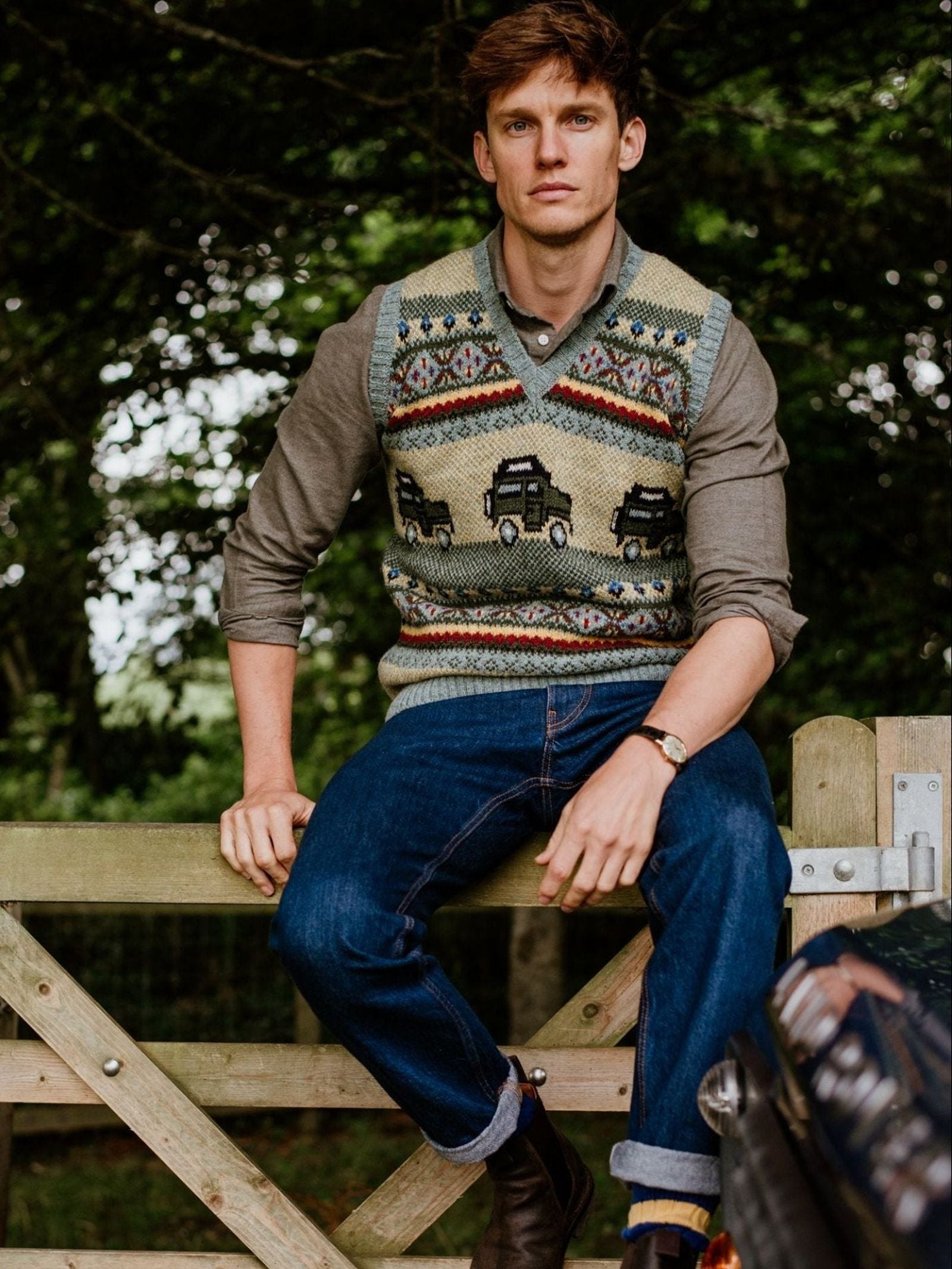 A man wearing the Campbell’s of Beauly Land Rover Defender Fairisle Slipover over a button-up shirt and jeans sits on a wooden fence outdoors, surrounded by green trees.