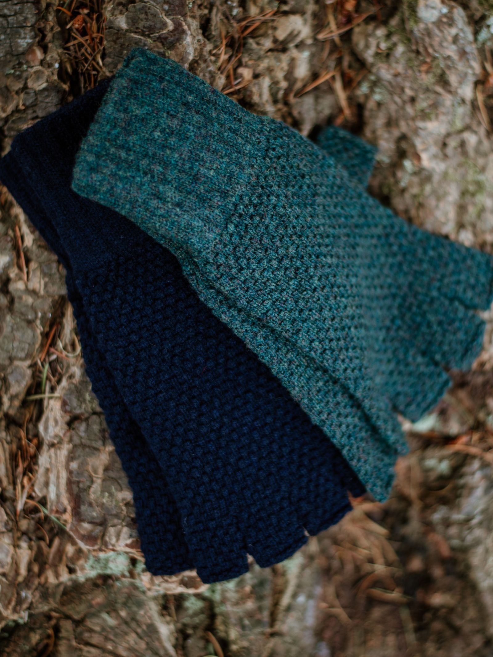 Two pairs of Campbell's of Beauly Moss Stitch Half-Finger Gloves, in dark blue and teal, rest on rough tree bark dotted with pine needles—an inviting winter essential.
