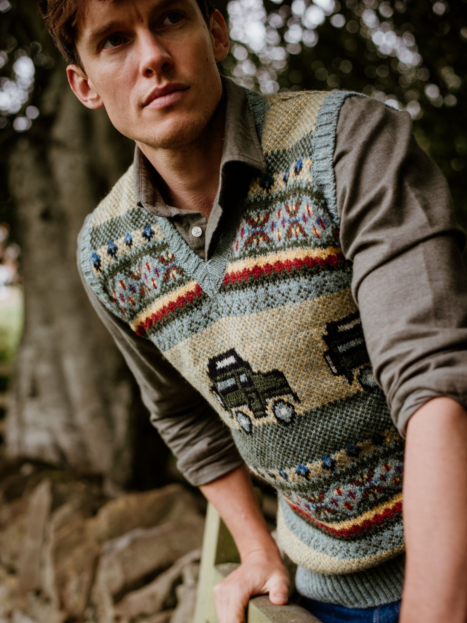 A young countryside enthusiast wears the Campbell's of Beauly Land Rover Defender Fairisle Slipover, featuring colorful patterns and black trucks, over a button-up shirt with trees and rocks blurred in the background.
