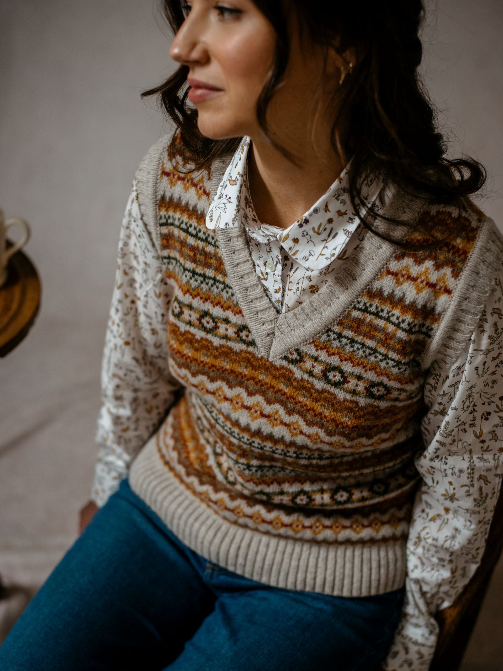A woman with wavy brown hair wears the Campbell's of Beauly Auld Stock Fairisle V-Neck Tank over a floral button-down shirt and blue jeans, seated on a chair and looking to the side—capturing timeless Scottish style.