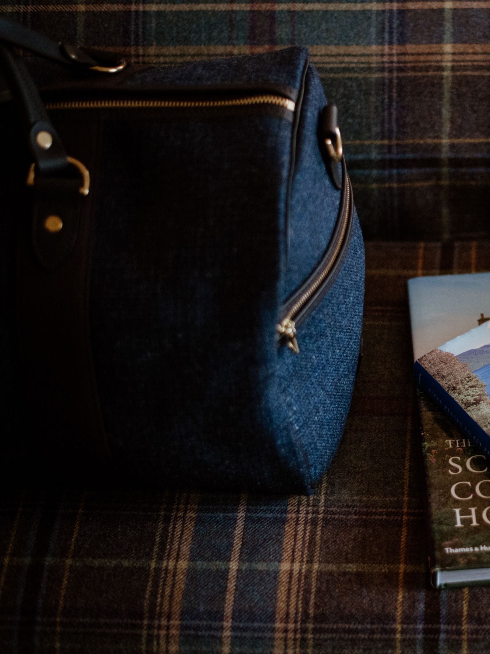 A Campbell's of Beauly Hand Luggage Holdall sits on a plaid-patterned couch next to a book with a scenic cover.