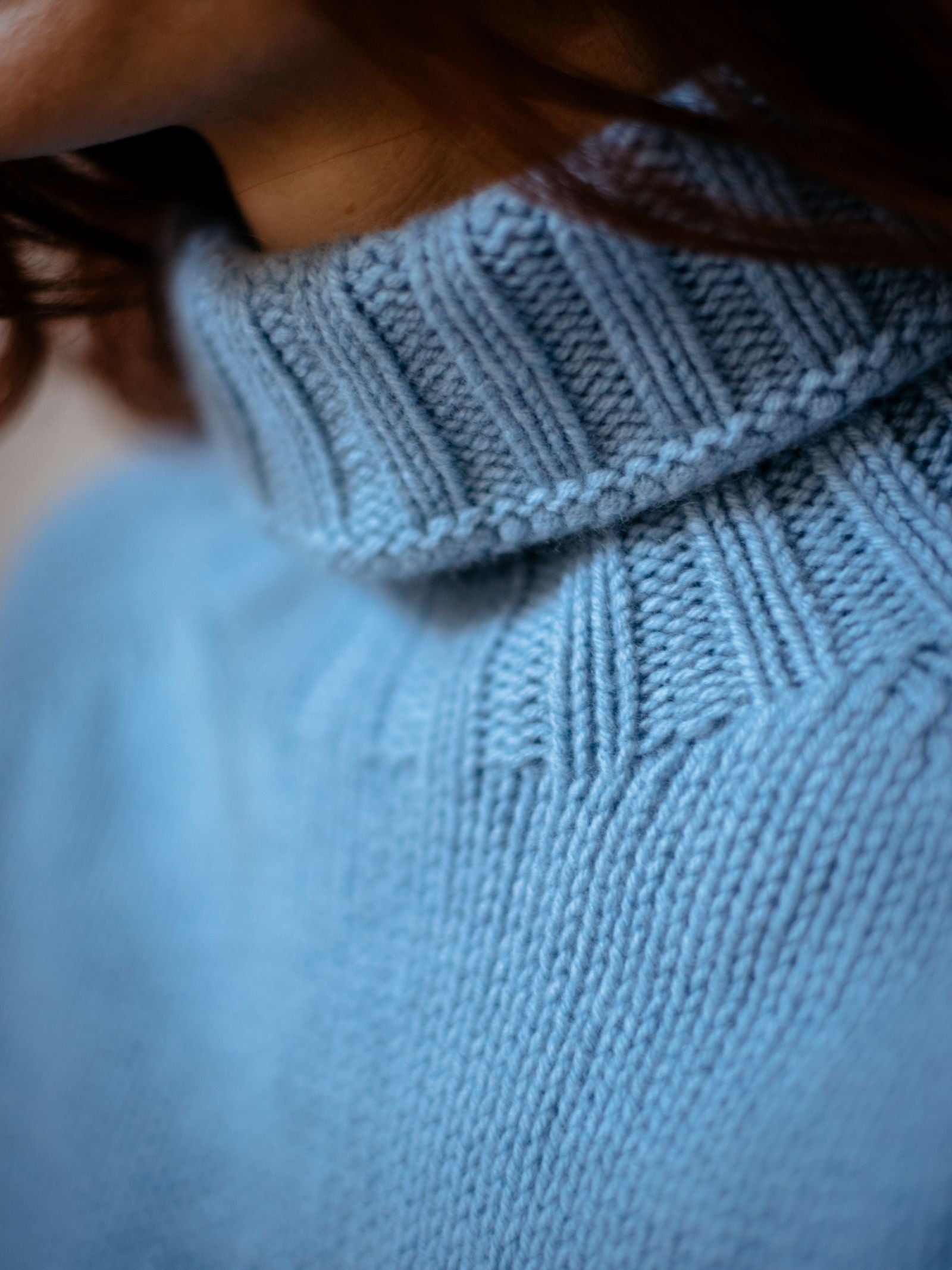 Close-up of a person wearing the Chunky Roll Neck Sweater by Campbell's of Beauly in light blue, with partially visible brown hair. The ribbed texture and supersoft wool of the sweater are clearly displayed.