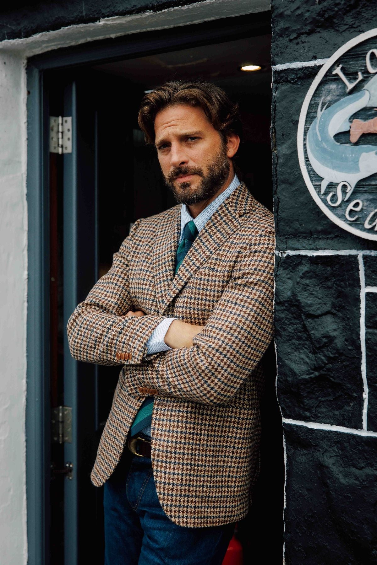 A man with brown hair and a beard stands in a doorway with arms crossed, wearing a checked blazer, blue shirt, green tie, and jeans. A round sign with a fish illustration is visible on the black wall beside him.