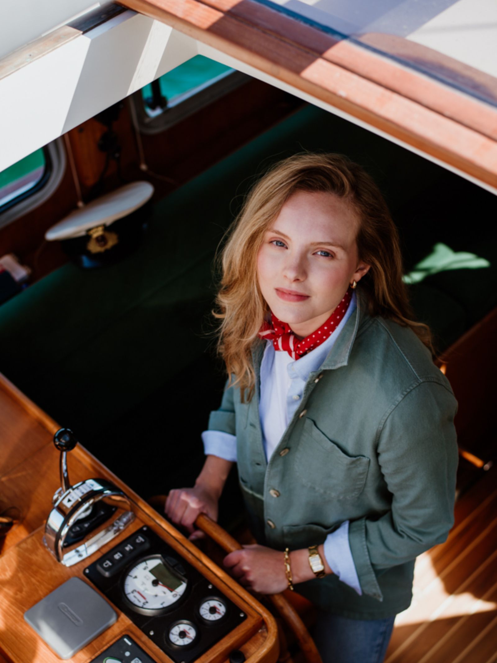 A woman with blonde hair wears the Campbell's of Beauly Auld Stock Summer Chore Jacket in green, standing at a boat’s helm. Sunlight pours in as nautical instruments and a captain’s hat rest nearby.