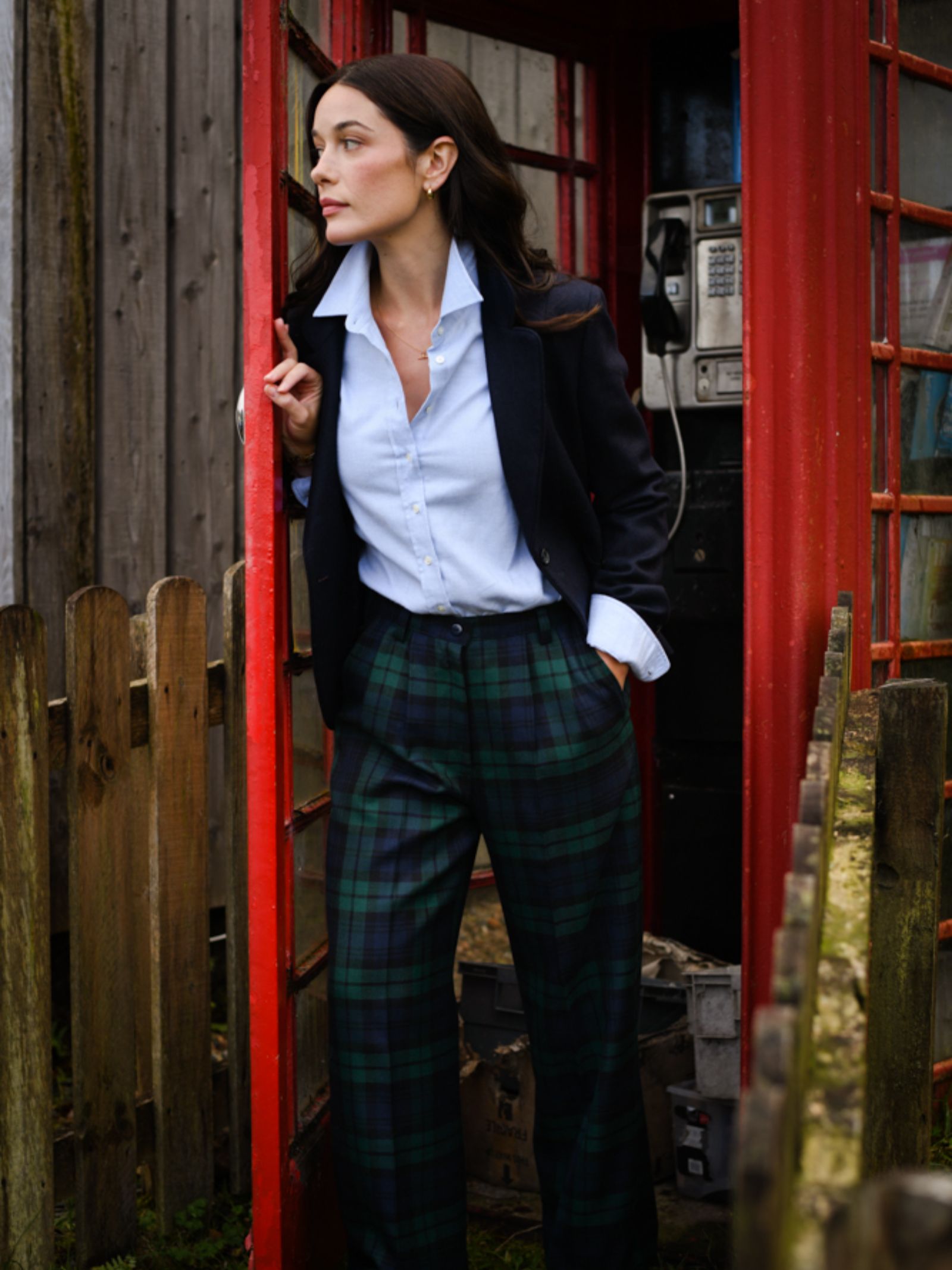A woman in a navy blazer, light blue shirt, and Campbell's of Beauly Wide Leg Blackwatch Trousers stands in a red British phone booth doorway, glancing to the side. A wooden fence and an old payphone are visible behind her.