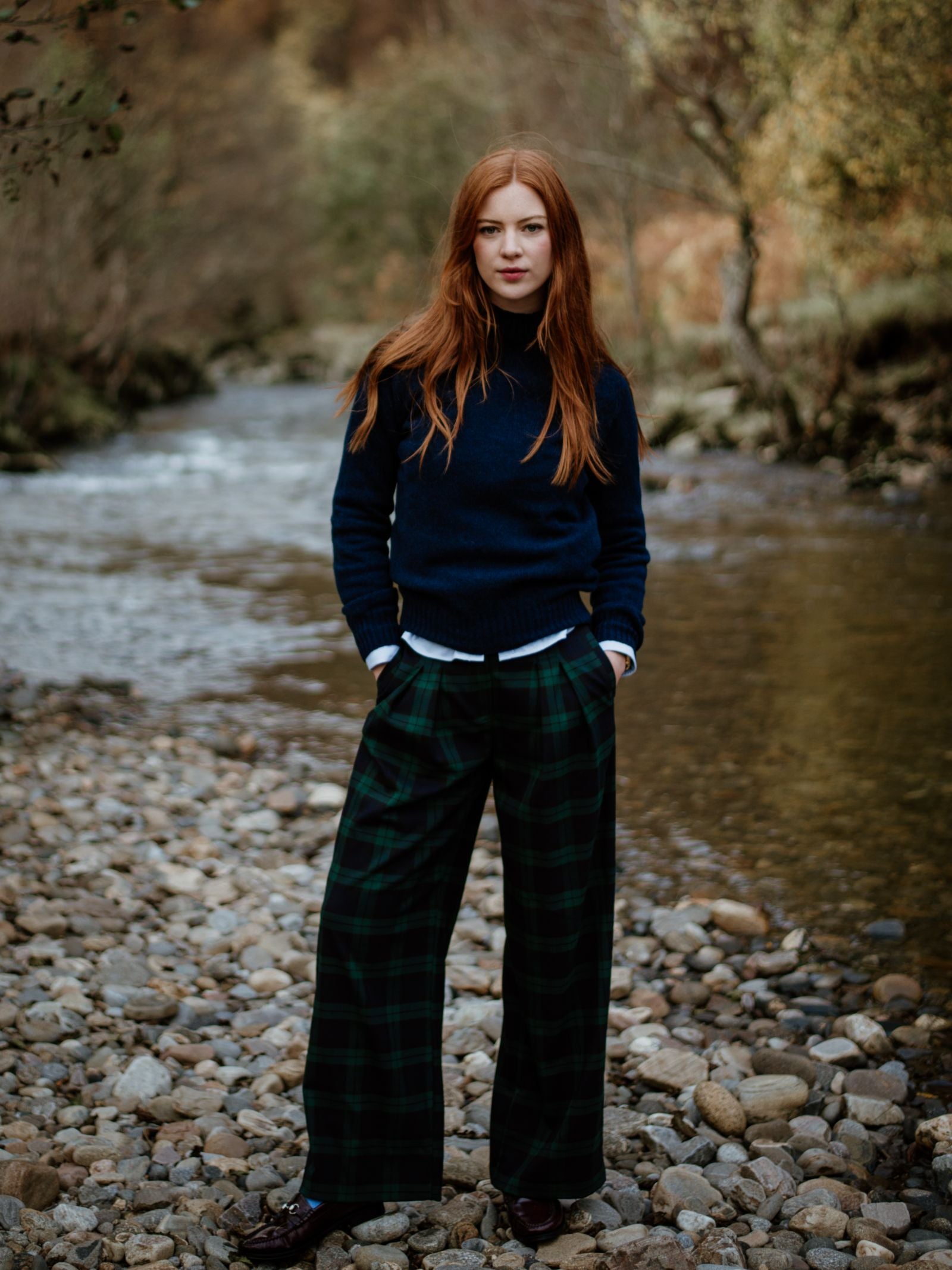 A woman with long red hair, wearing a Campbells of Beauly Geelong Lambswool Funnel Neck and green plaid wide-leg pants, stands on a rocky riverbank with water and trees in the background.