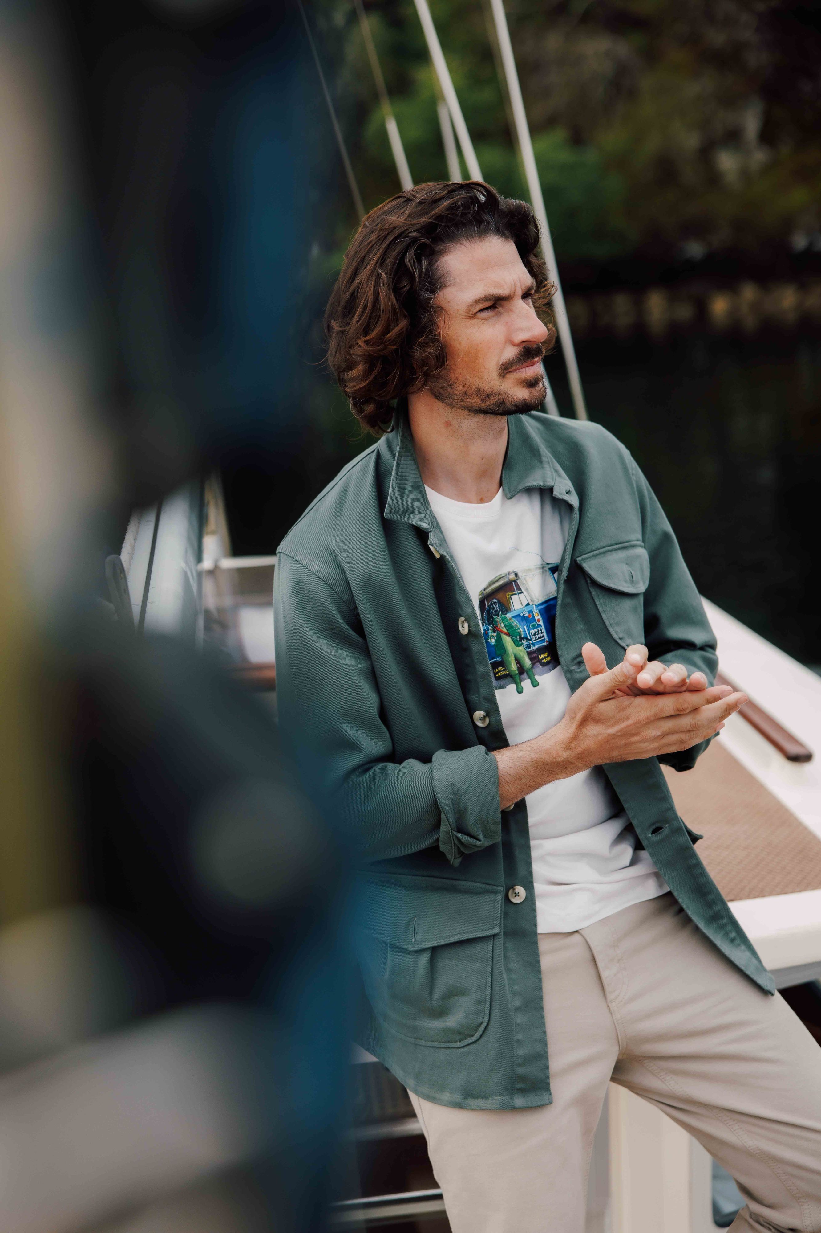 A man with wavy brown hair and facial hair leans against a boat railing, wearing a green jacket over a graphic T-shirt and beige pants. He looks thoughtfully into the distance, with trees and water in the background.