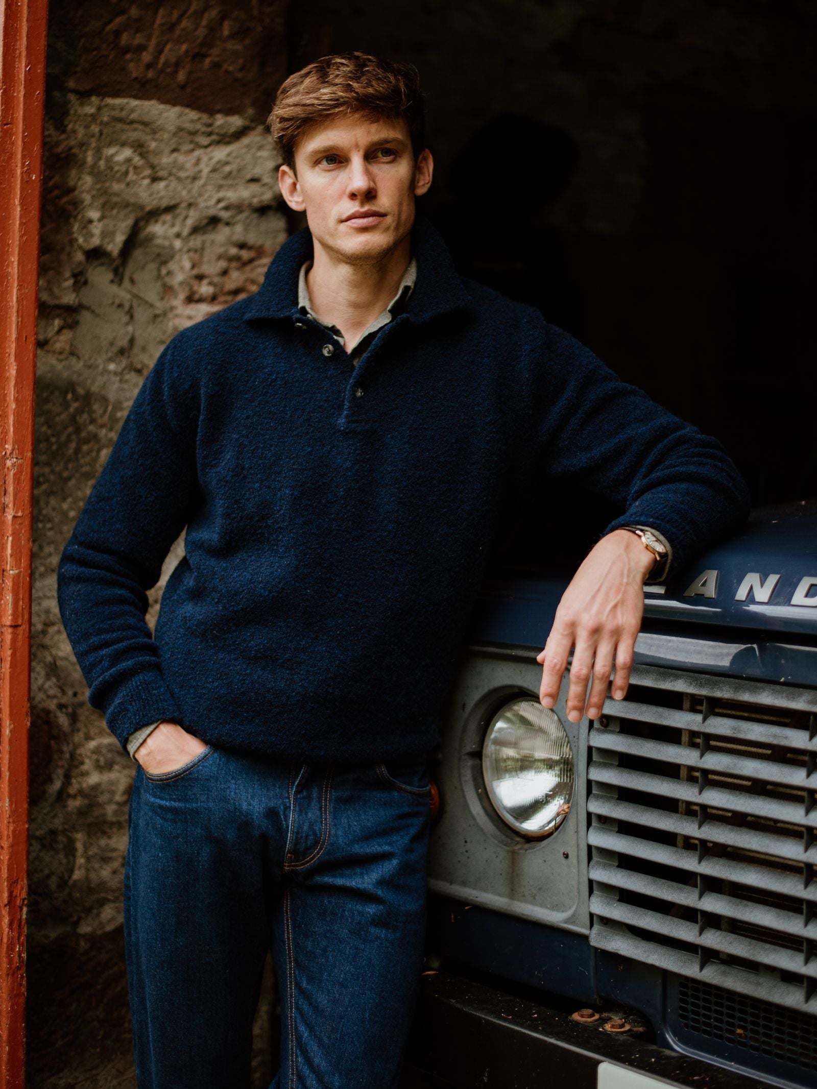 A man with short brown hair, wearing the Campbell's of Beauly Auld Stock Bouclé Neck Jumper and jeans, stands by a stone wall with one hand in his pocket and the other on a vintage blue Land Rover, looking serious.