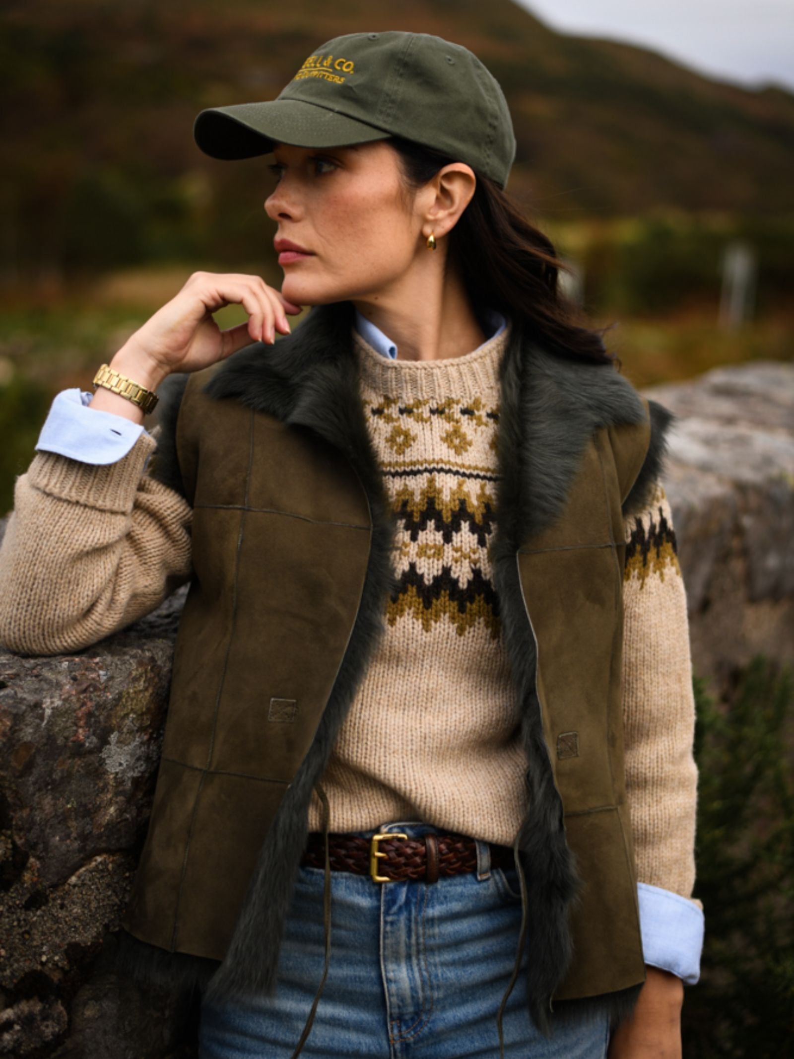 A woman in a green cap, patterned sweater, and jeans leans against a stone wall outdoors, wearing the Campbell's of Beauly Auld Stock Sheepskin Reversible Gilet. Greenery and hills fill the background as she looks thoughtfully aside.