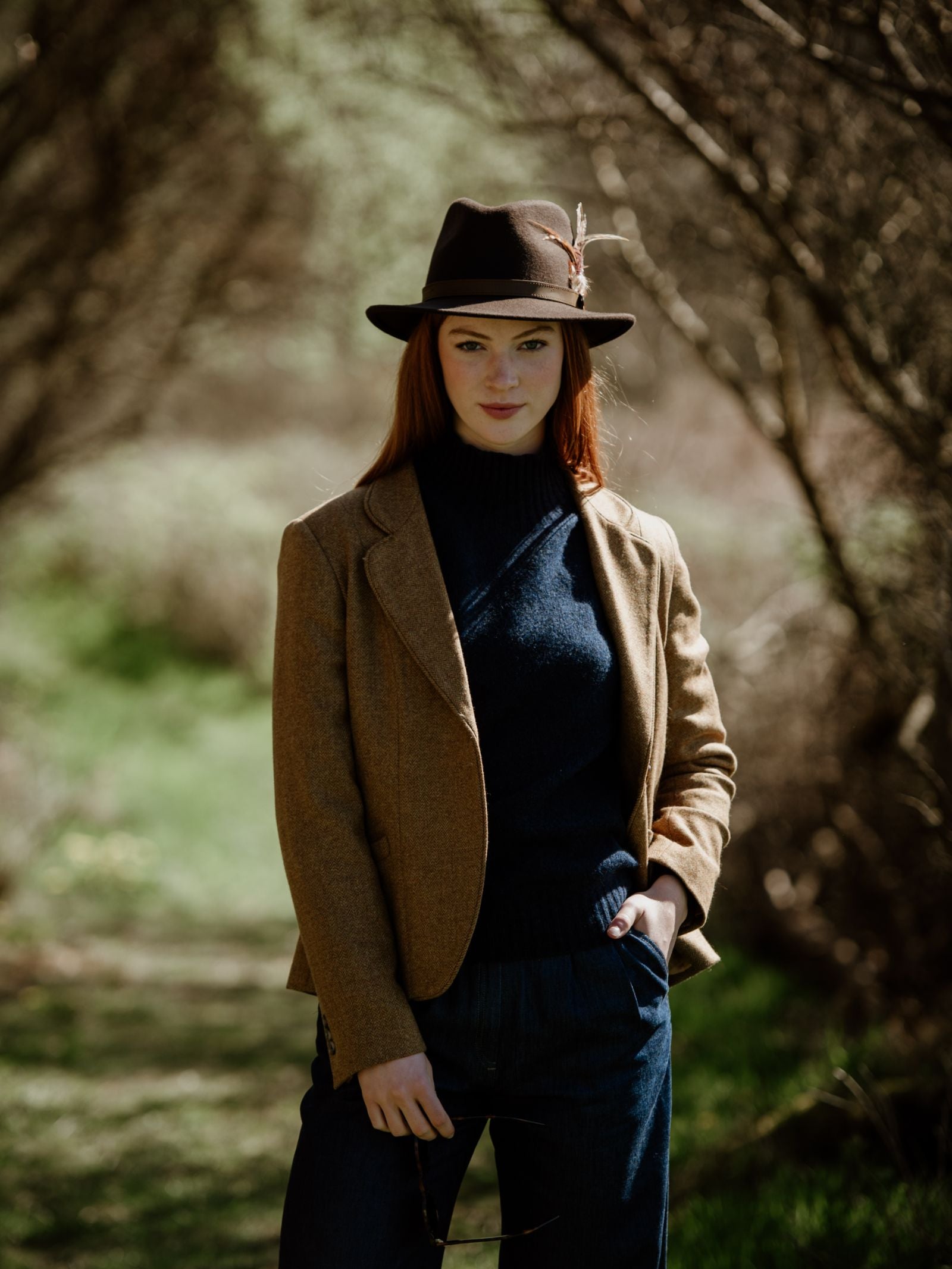 A woman with long red hair stands outdoors on a grassy path, wearing the Hicks & Brown Auld Stock Gamebird Fedora with feather wrap, a tan blazer, dark sweater, and jeans. Sunlight filters through trees in the dappled background.