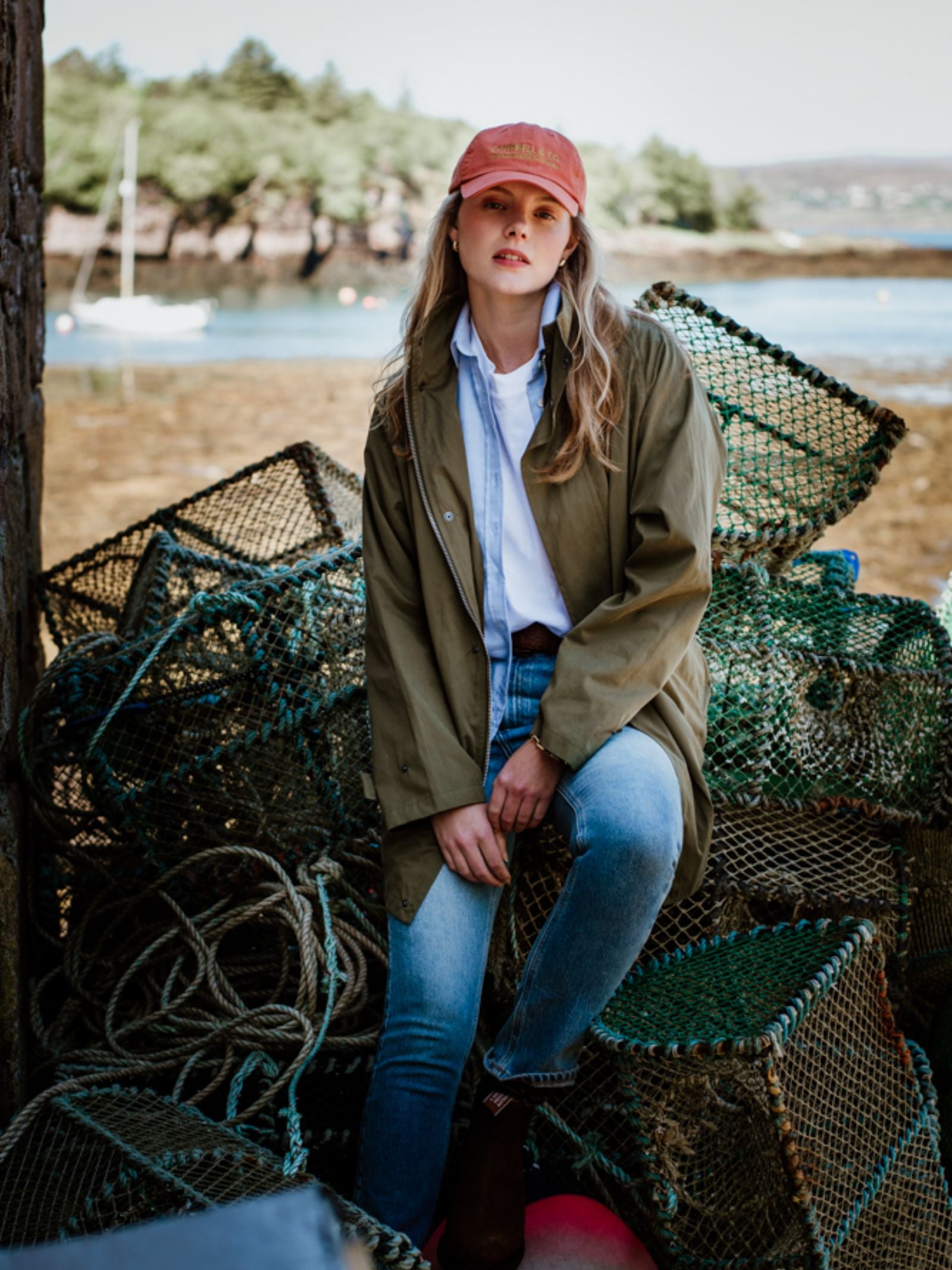 A woman in a Unisex Campbell's Baseball Cap by Campbell's of Beauly, green raincoat, white shirt, and blue jeans sits on stacked fishing traps by the water, with trees and a boat in the background.