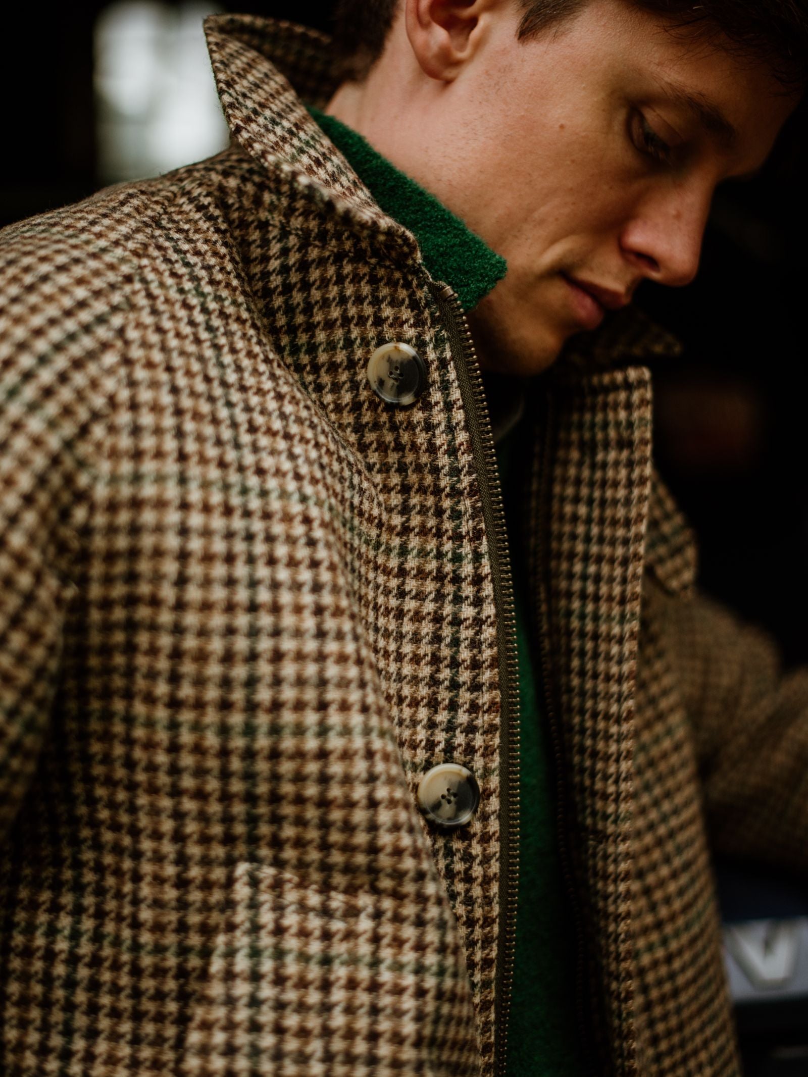 A person wearing the Cannich Coat by Campbell's of Beauly—a brown and cream houndstooth wool coat with large buttons—over a green sweater, looking downward in soft lighting.