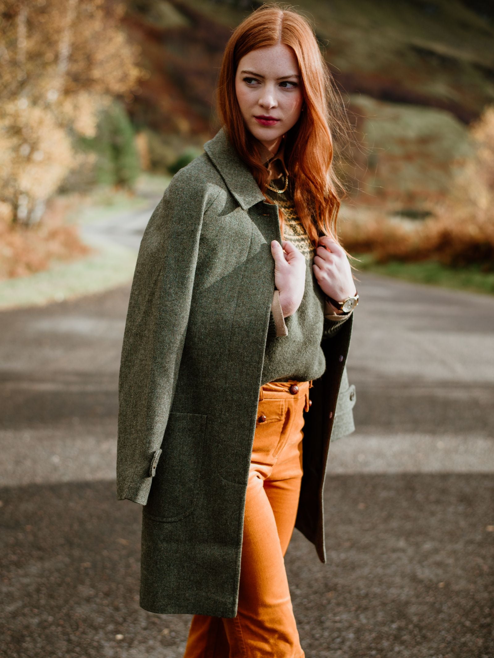 A woman with long red hair wears the knee-length olive green Auld Stock Heath Collar Coat by Campbell's of Beauly and orange pants on a paved path, surrounded by autumn trees—an ideal choice for stylish everyday wear.