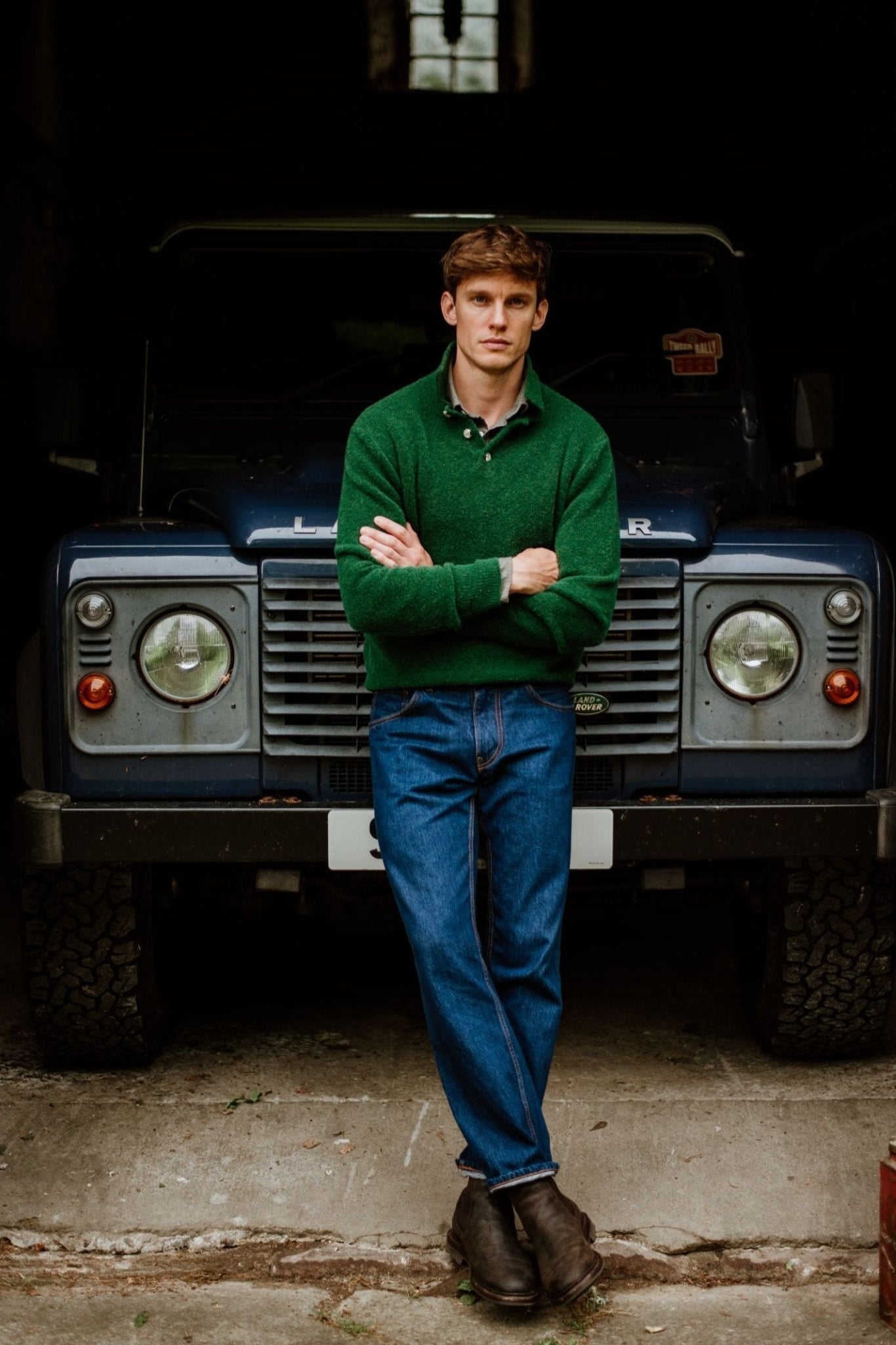 A man in a green Auld Stock Bouclé Neck Jumper by Campbell's of Beauly, blue jeans, and brown boots stands with arms crossed before a blue Land Rover in a dimly lit garage.