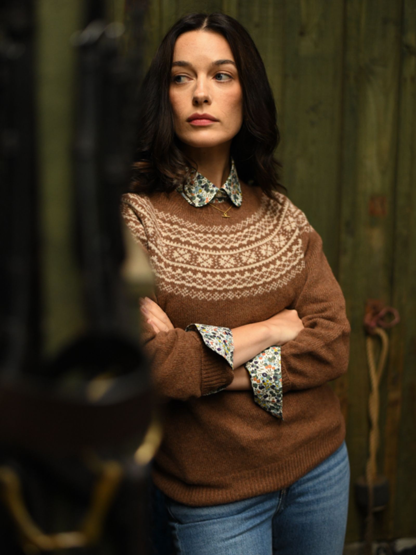 A dark-haired woman stands with arms crossed by a green wooden wall, wearing Campbell's of Beauly Auld Stock Two-Colour Fairisle Yoke Jumper over a floral collared shirt and blue jeans, looking to the side.
