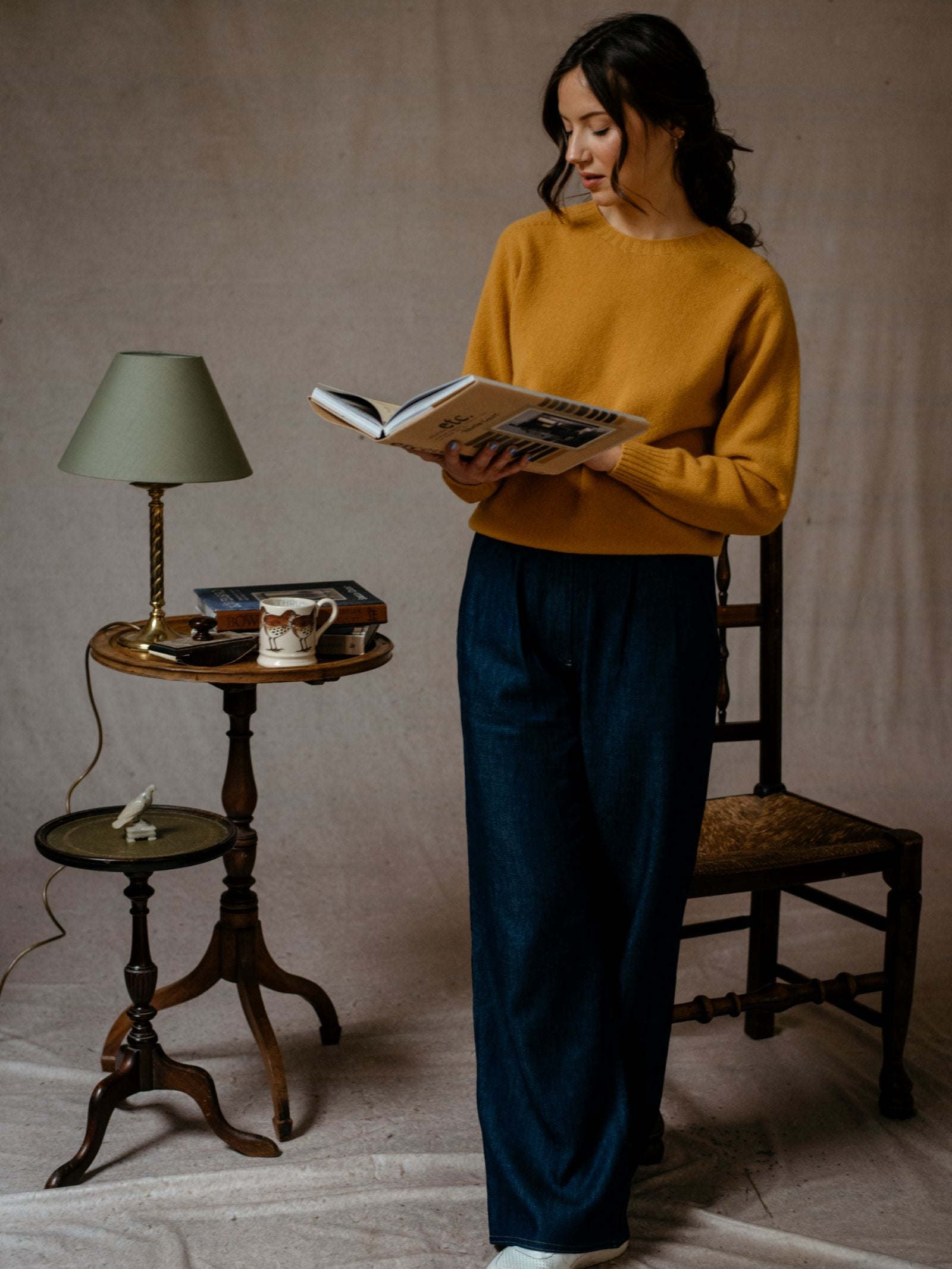 A woman in a mustard yellow Auld Stock Geelong Lambswool Crew Neck Jumper by Campbell's of Beauly and wide-leg jeans stands beside a wooden side table with a green lamp, books, and a cat-themed mug, reading in a cozy, softly lit room.