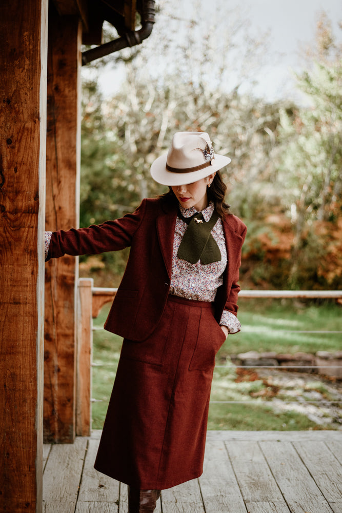 A woman in a vintage-style maroon suit, floral blouse, and wide-brimmed hat stands on a wooden porch, leaning against a post, with trees and grass visible in the background.