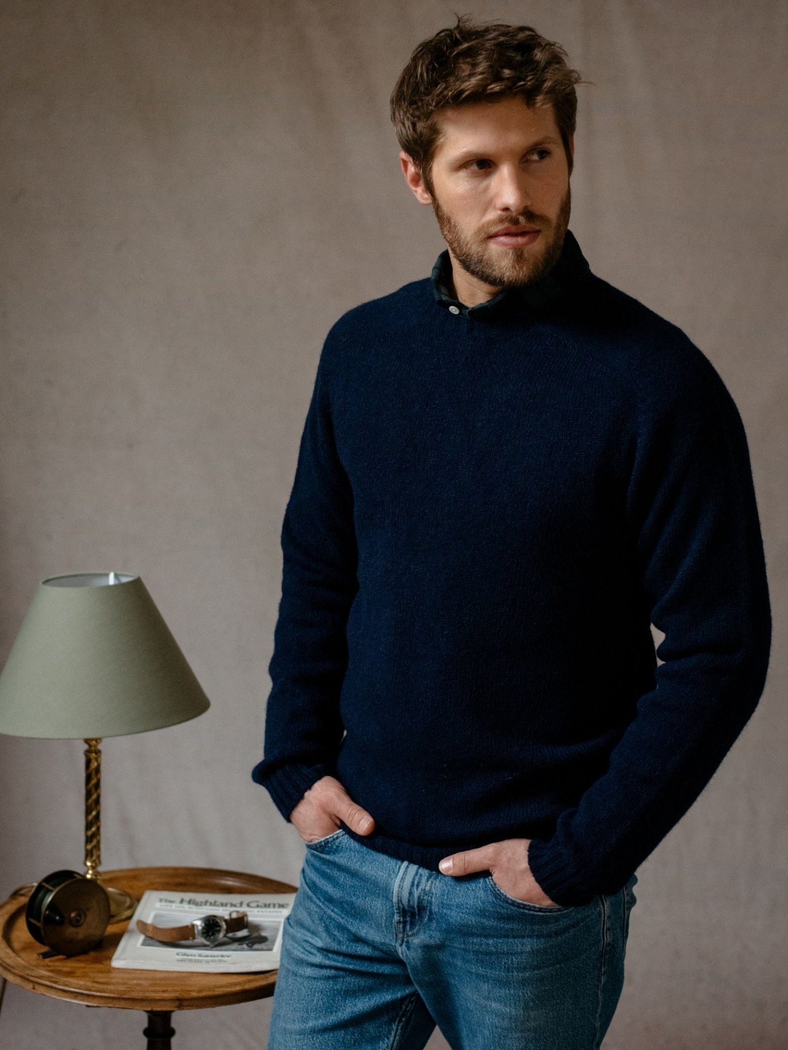 A man with short brown hair and a beard, wearing a dark blue Campbell's of Beauly Shetland Jumper and jeans, stands with hands in his pockets by a small round table holding a green lamp, glasses, and a magazine.
