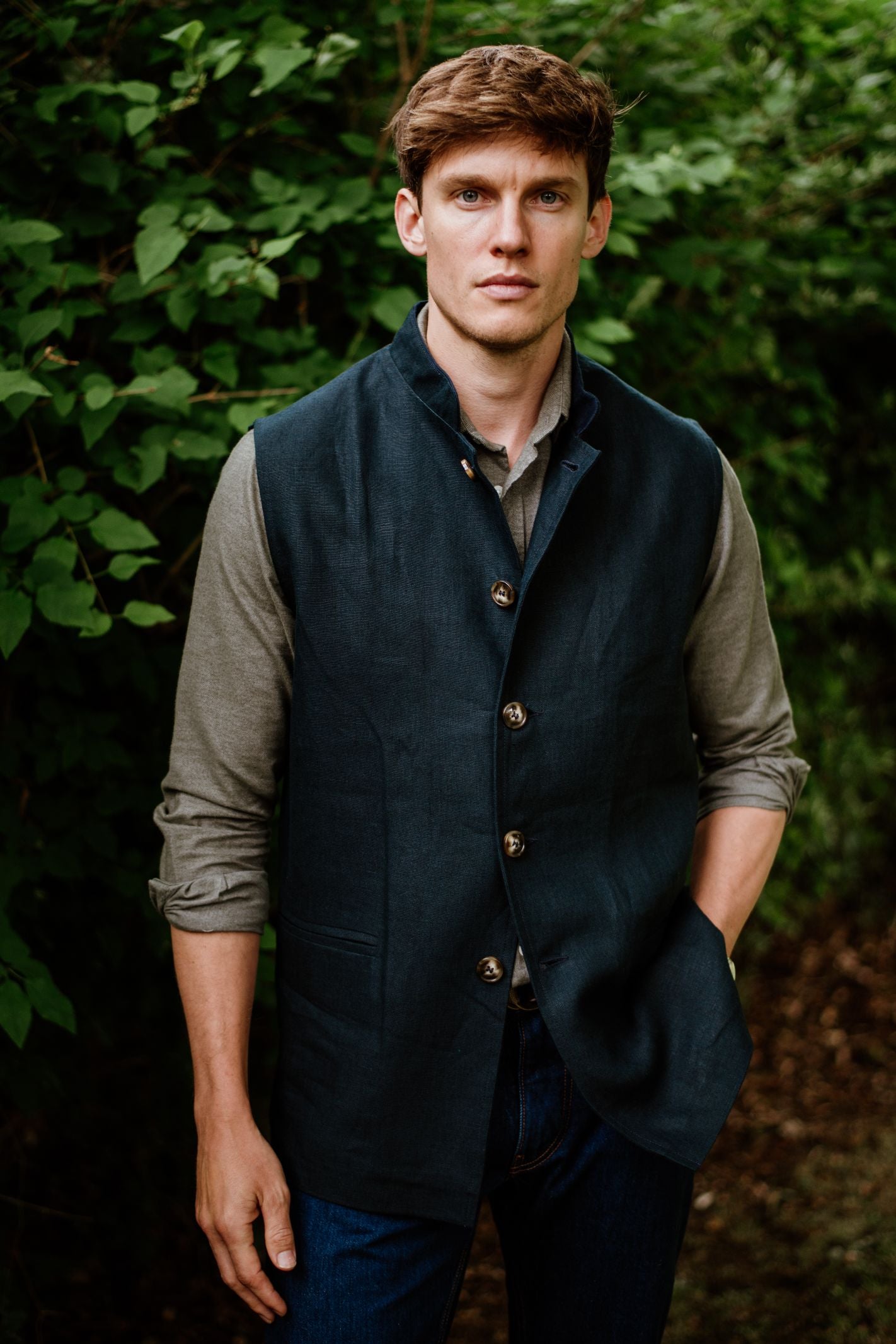 A man with short brown hair stands outdoors near greenery, wearing a Campbell's of Beauly Linen Nehru Gilet over a light olive shirt and dark jeans, one hand in his pocket as he looks at the camera.