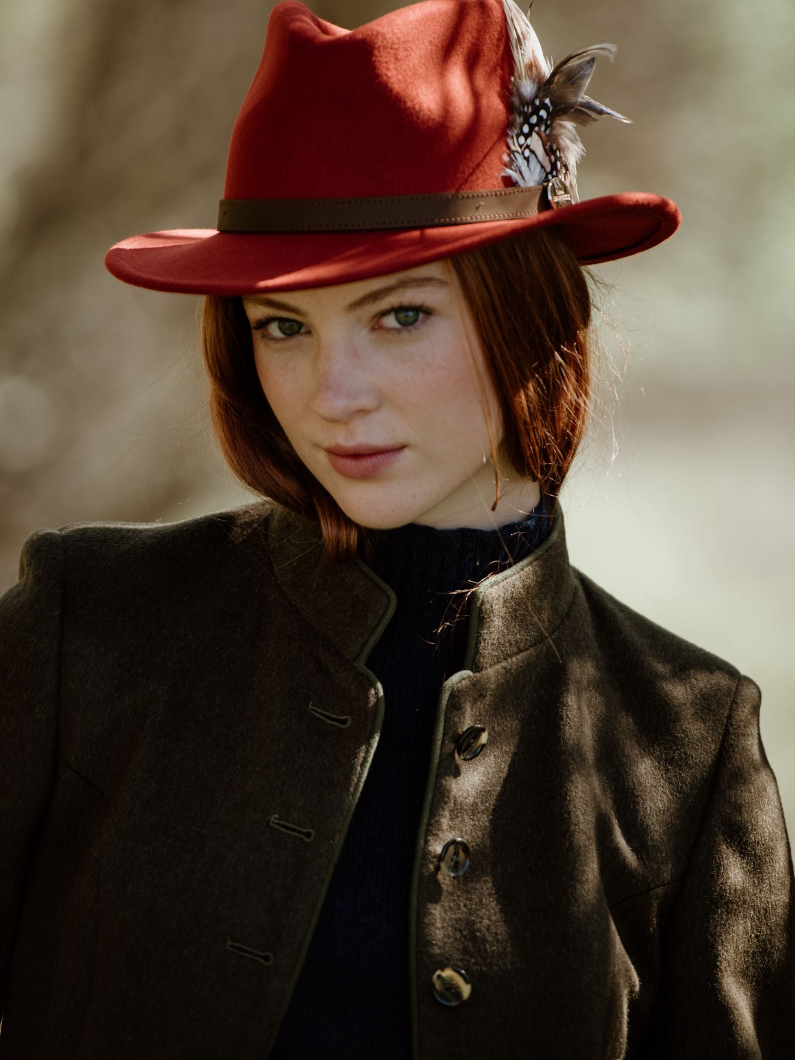 A young woman with fair skin and red hair wears the Campbells of Beauly Loden Green Millie Coat and a feathered red hat, looking softly at the camera against a blurred background.