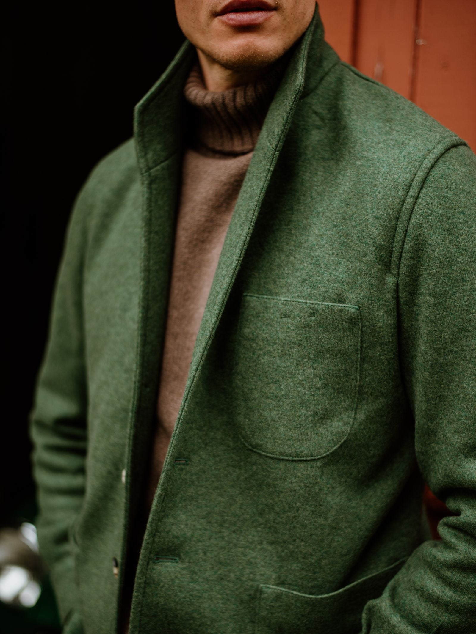 A man in a green Campbell's of Beauly Cashmere Teba Jacket over a brown turtleneck stands before a reddish-brown background, his partially visible face evoking timeless menswear style.