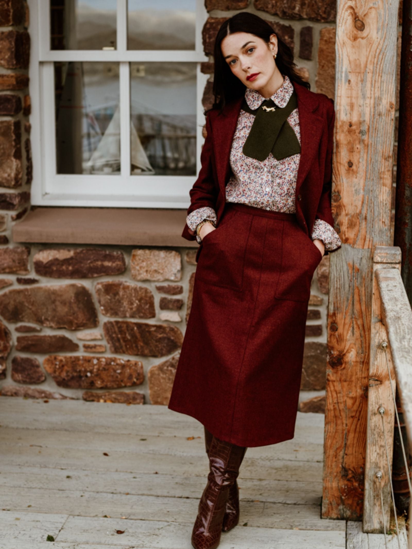 A woman leans on a rustic wooden porch, wearing the Campbell's of Beauly Auld Stock A-Line Skirt with a matching blazer, floral bow blouse, and knee-high brown boots. A stone wall and window appear in the background.