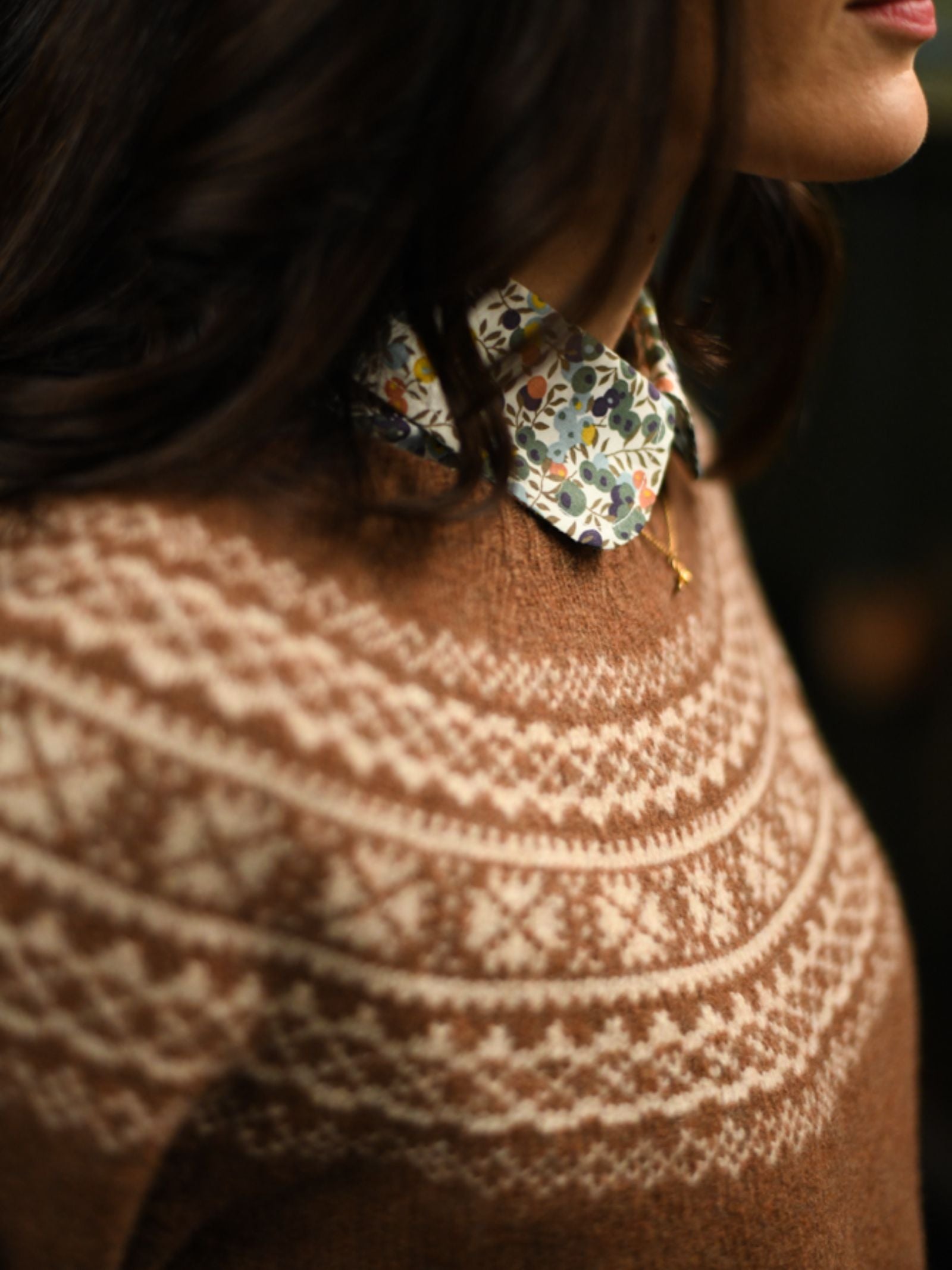 A woman wears the Campbell's of Beauly Auld Stock Two-Colour Fairisle Yoke Jumper, featuring a brown base with a white Nordic pattern, layered over a floral collared shirt. Only her torso and lower face are shown.