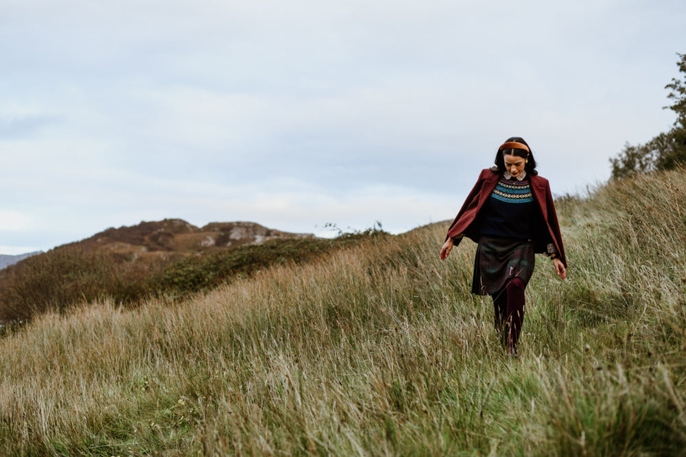 A woman in a red coat and skirt walks through tall grass on a hillside, with rolling hills and an overcast sky in the background.