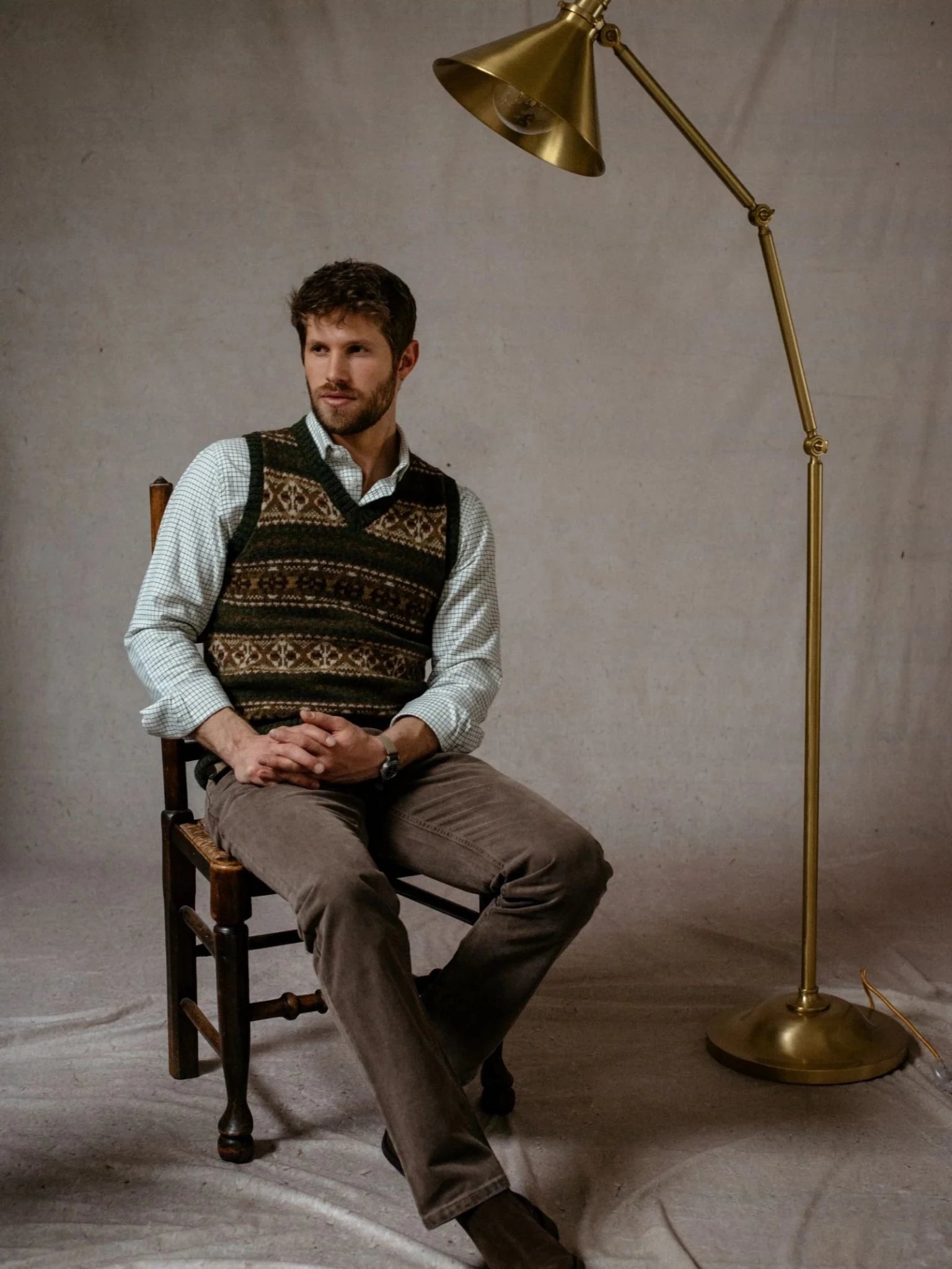 A man wearing the Campbell’s of Beauly Fairisle Slipover—sleeveless knitwear crafted from genuine Shetland wool—sits on a wooden chair beside a brass floor lamp, set against a plain backdrop.