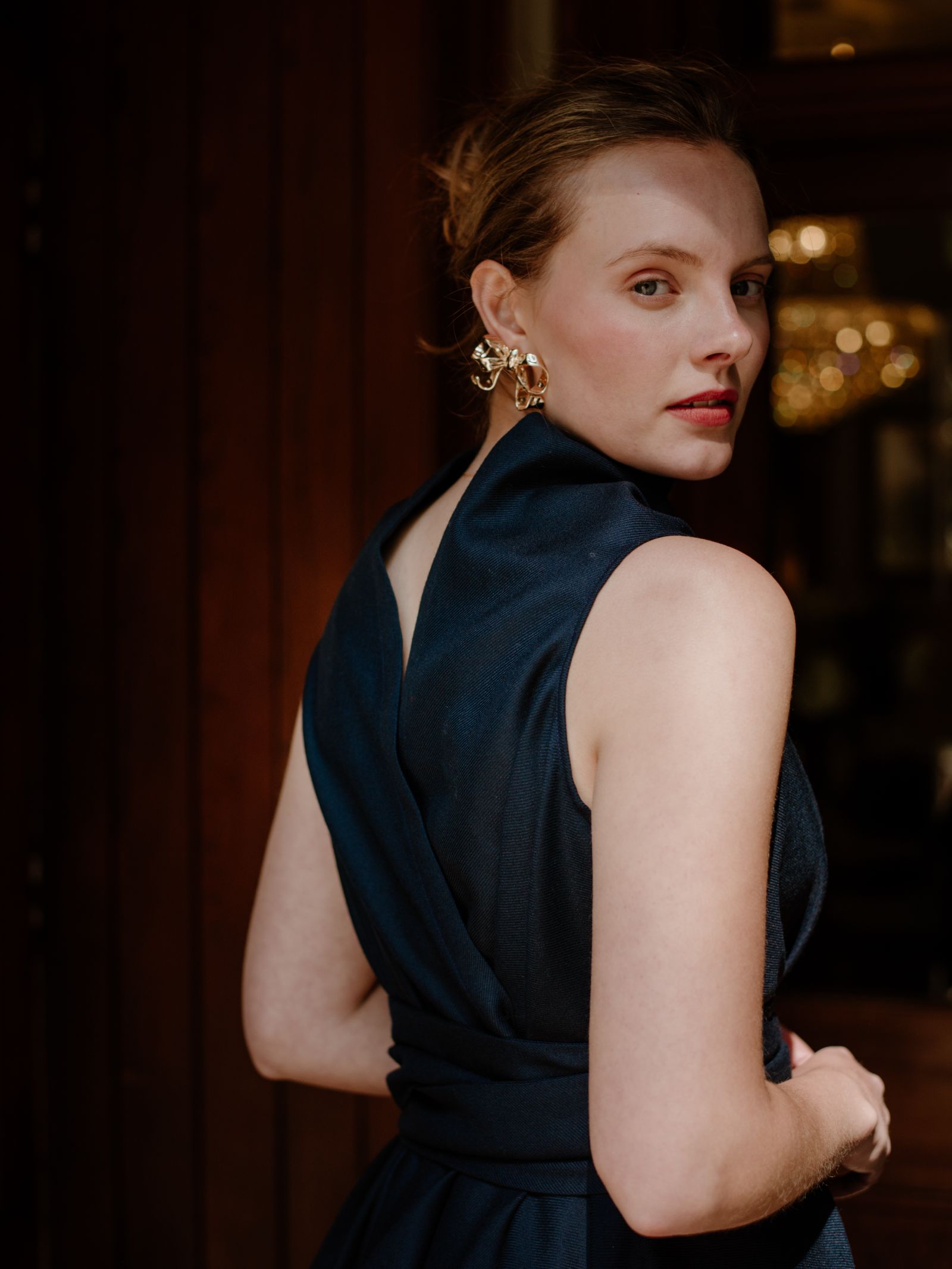 A woman in a dark blue, reversible wool wrap dress by Campbell's of Beauly stands with her back turned, looking over her shoulder. She wears gold statement earrings and light makeup, set against a softly lit indoor background.