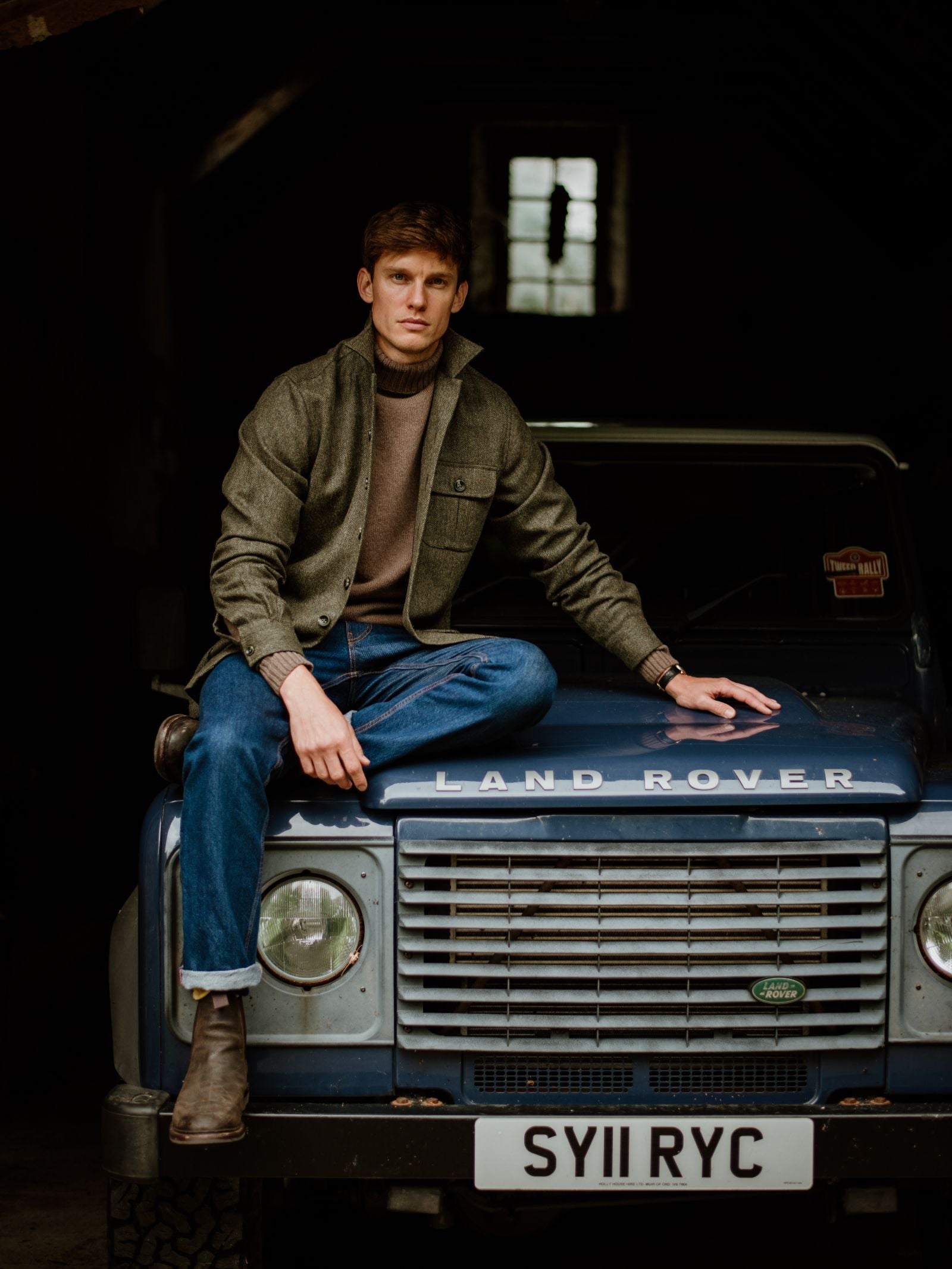 A man in a Campbell's of Beauly Casual Tweed Shacket and blue jeans sits on the hood of a blue Land Rover Defender (SY11 RYC) in a dimly lit garage, with light streaming through the window behind him.