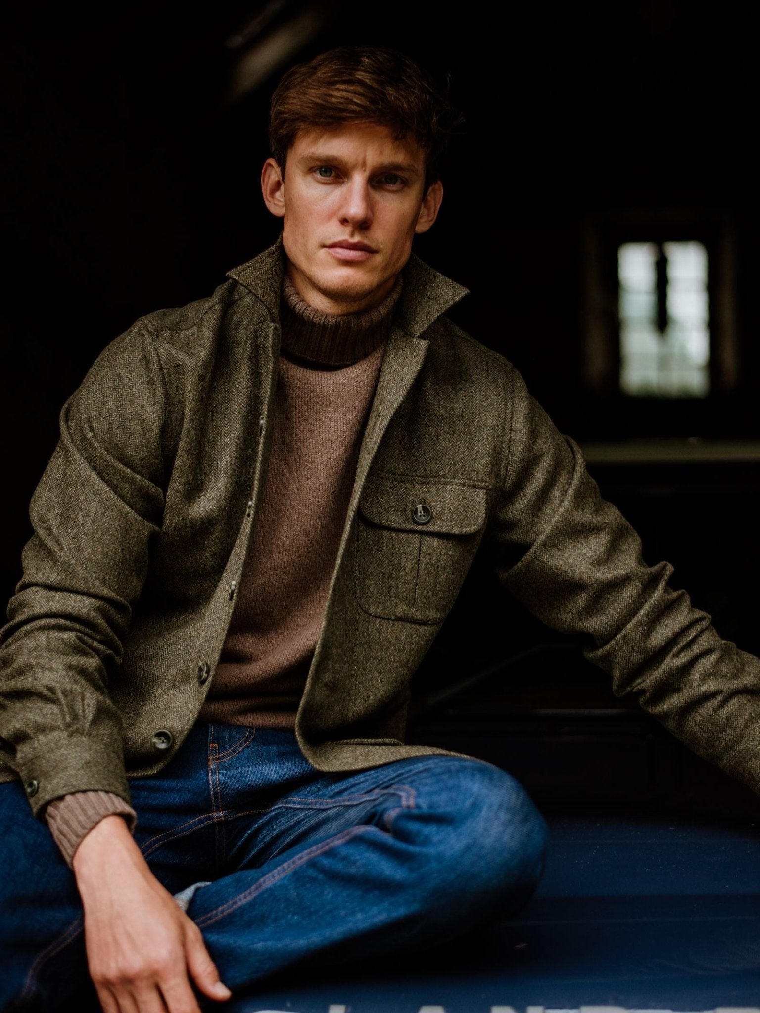 A man with short brown hair sits casually, one knee up, wearing the Campbell's of Beauly Casual Tweed Shacket over a brown sweater and blue jeans. Soft light from a window behind him highlights his relaxed style against a dark background.