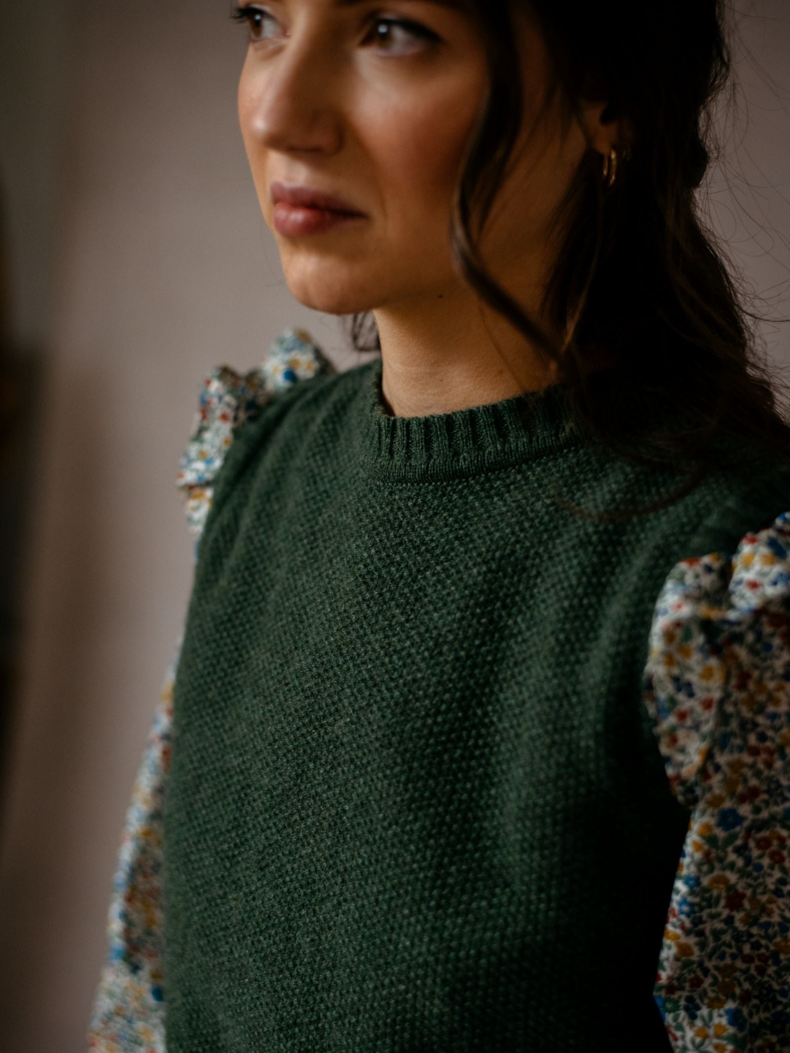 A woman with long dark hair wears the Moss Stitch Crew Tank by Campbells of Beauly, featuring textured Scottish yarn and floral-patterned puffy sleeves. She looks slightly to the side, shown from the shoulders up against a neutral background.