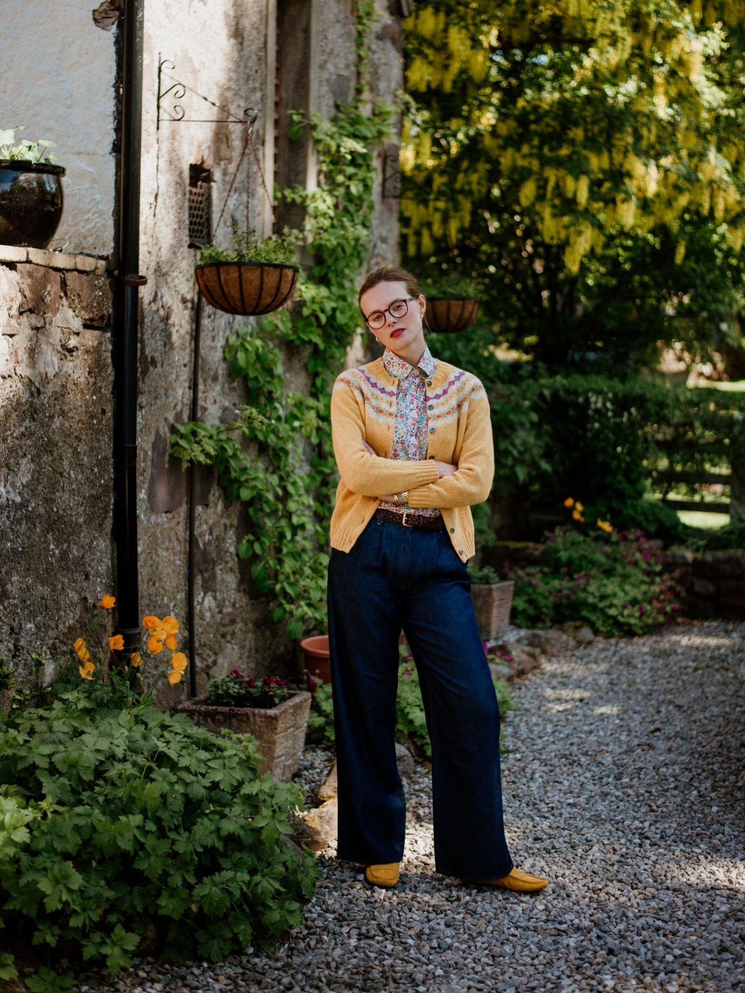 Standing outside by a stone wall with greenery and flowers, the person wears glasses, a yellow cardigan, patterned shirt, tie, Campbell's of Beauly Auld Stock Wide Leg Jeans in lightweight denim, yellow shoes, arms crossed and neutral expression.