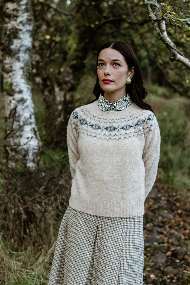 A woman with long dark hair stands outdoors among trees, wearing a light wool sweater with a patterned yoke, a floral-collared shirt, and a plaid skirt. She gazes slightly upward with a calm expression.