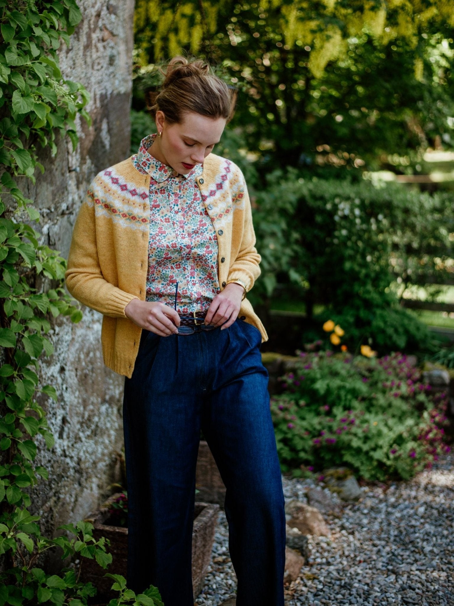 A woman stands outdoors by a leafy stone wall, wearing Campbell's of Beauly Shetland Fairisle Cardigan in yellow over a floral blouse and dark blue high-waisted trousers, with lush gardens and trees behind her.