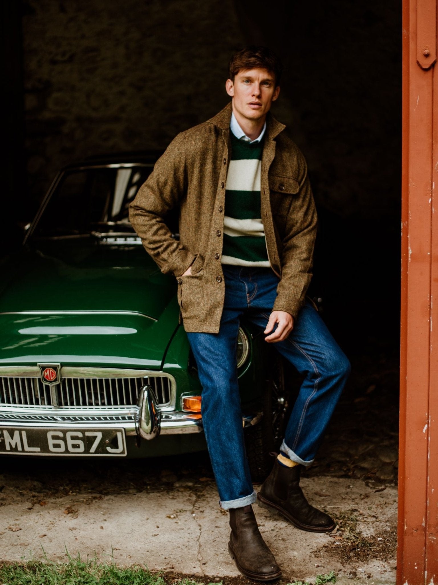A man in a Campbell's of Beauly Casual Tweed Shacket, striped sweater, blue jeans, and brown boots sits on the bumper of a classic green MG car inside a dimly lit garage.