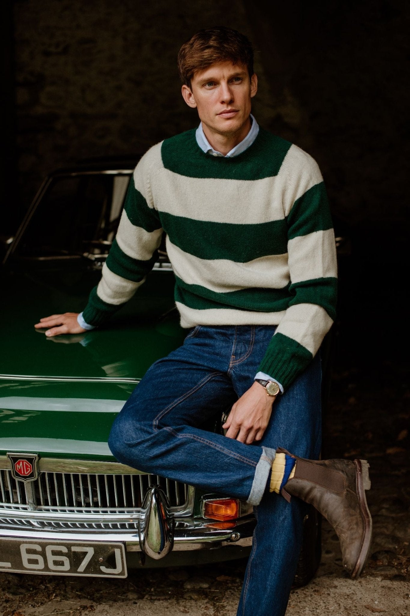 A man in a Campbell's of Beauly Auld Stock Shetland Stripe Crew Jumper, blue jeans, and brown boots sits on the hood of a classic green MG car, gazing to the side with a neutral expression.