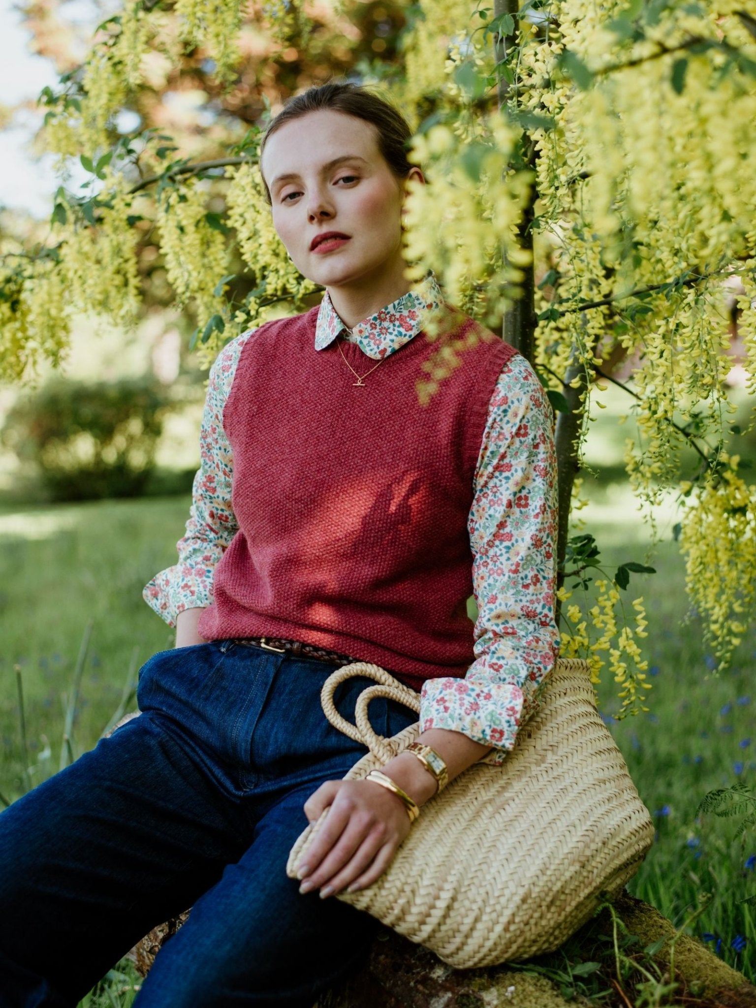 A woman sits outdoors on a rock, wearing blue jeans and the Campbell’s of Beauly Auld Stock Moss Stitch Crew Tank over a fine gauge floral shirt, holding a straw bag. She is surrounded by greenery and yellow flowers, looking calm and confident.