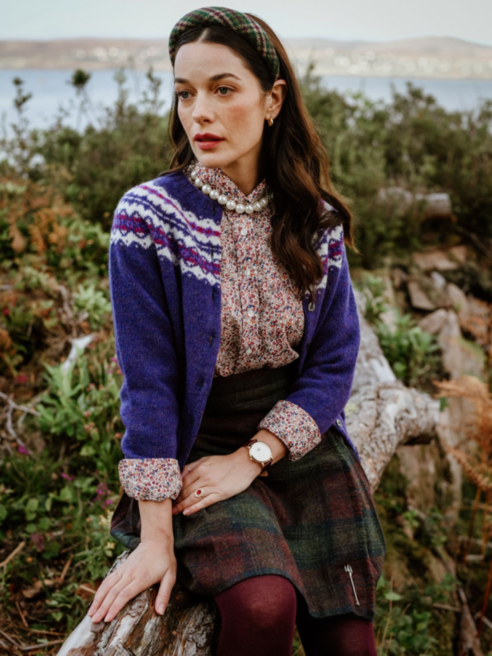 A woman in a patterned cardigan, floral blouse, and plaid skirt sits on a log outdoors, wearing a headband and watch, with greenery and water in the background.