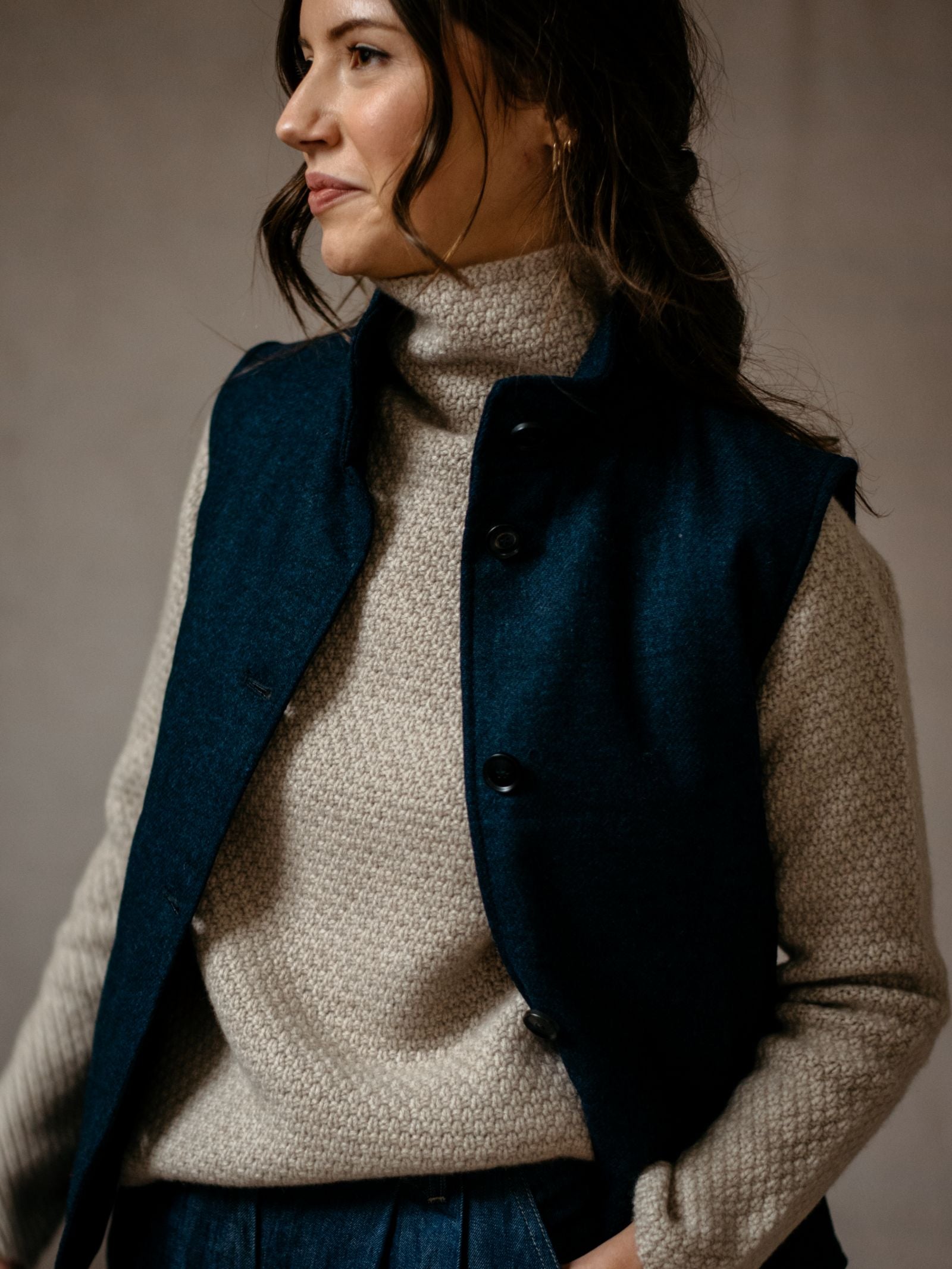 A woman with wavy brown hair wears a textured beige turtleneck and the Campbell’s of Beauly Auld Stock Quilted Buttoned Gilet in dark blue tweed, posing with her hand in her pocket—a stylish layering piece for any season.