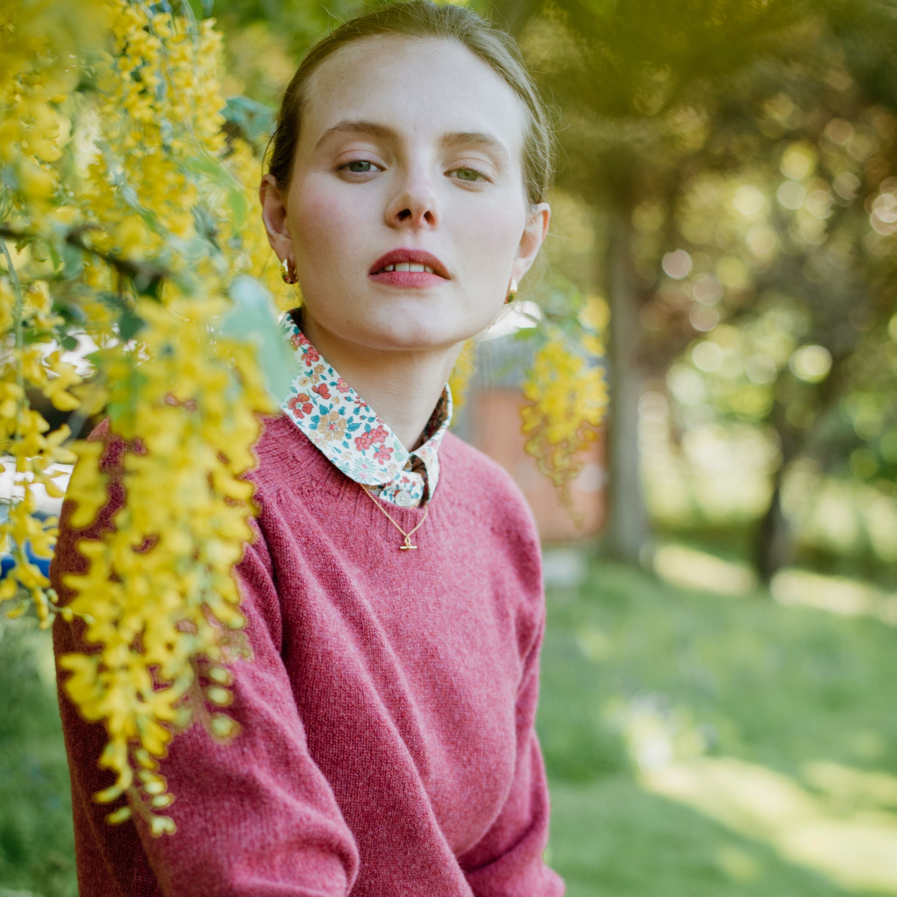 A woman wearing a pink sweater and a floral-collared shirt poses outdoors next to hanging clusters of yellow flowers, with sunlight filtering through green foliage in the background.
