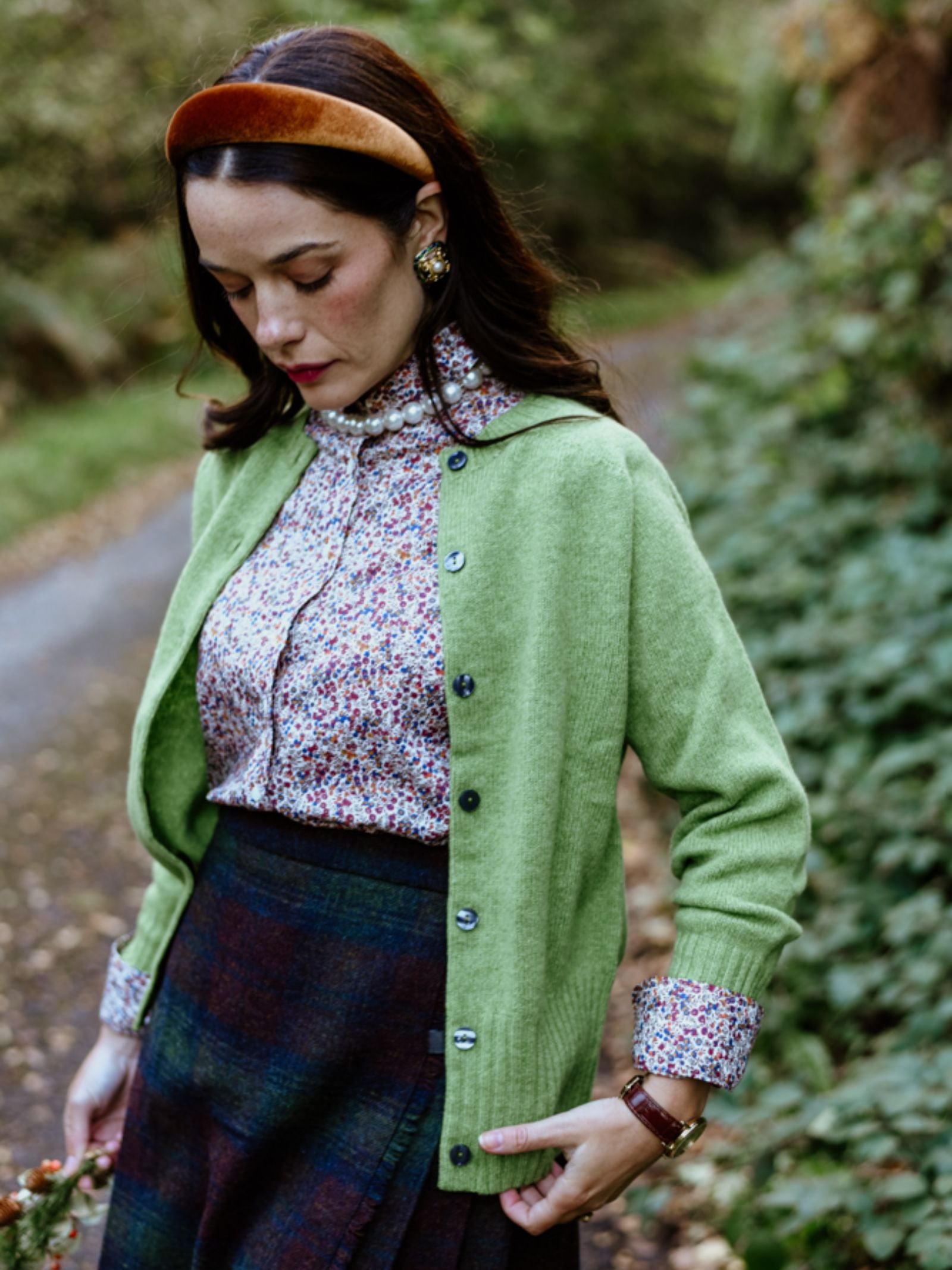 A woman with dark hair stands on a leafy path wearing Campbell's of Beauly Geelong Lambswool Crew Cardigan in green over a floral blouse and plaid skirt, accessorized with a brown headband and gold earrings.