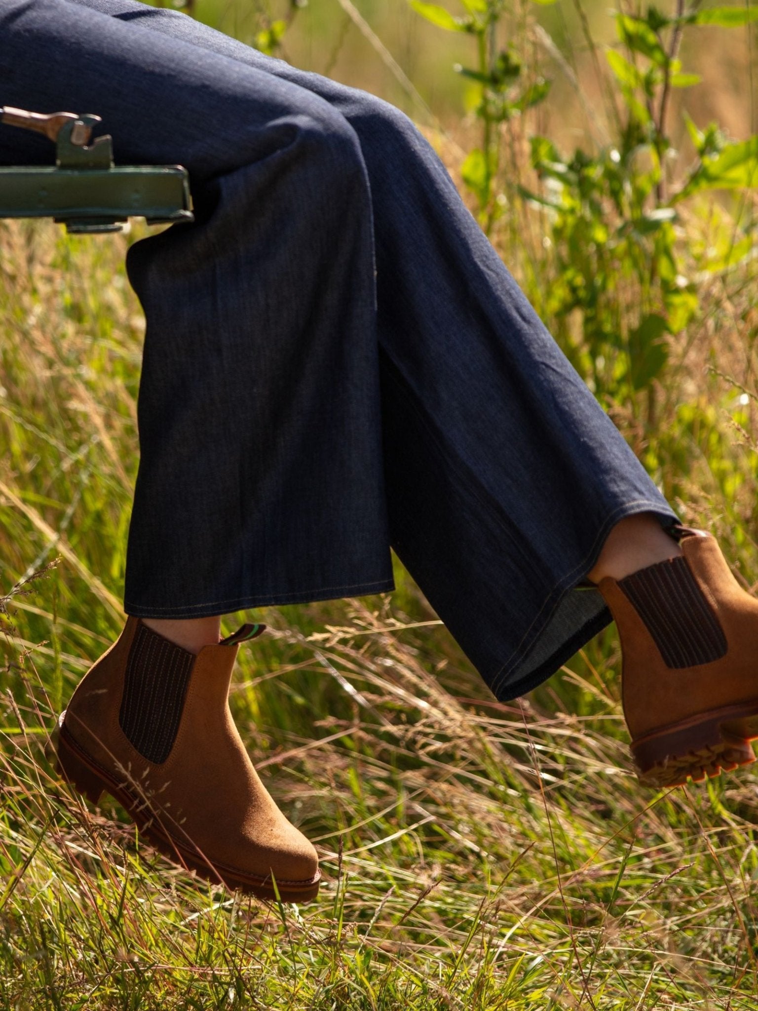Relaxing outdoors amid tall grass, a person crosses their legs in wide-leg blue jeans and Leather Chelsea Boots by Campbell's of Beauly, creating a charming country-inspired look.