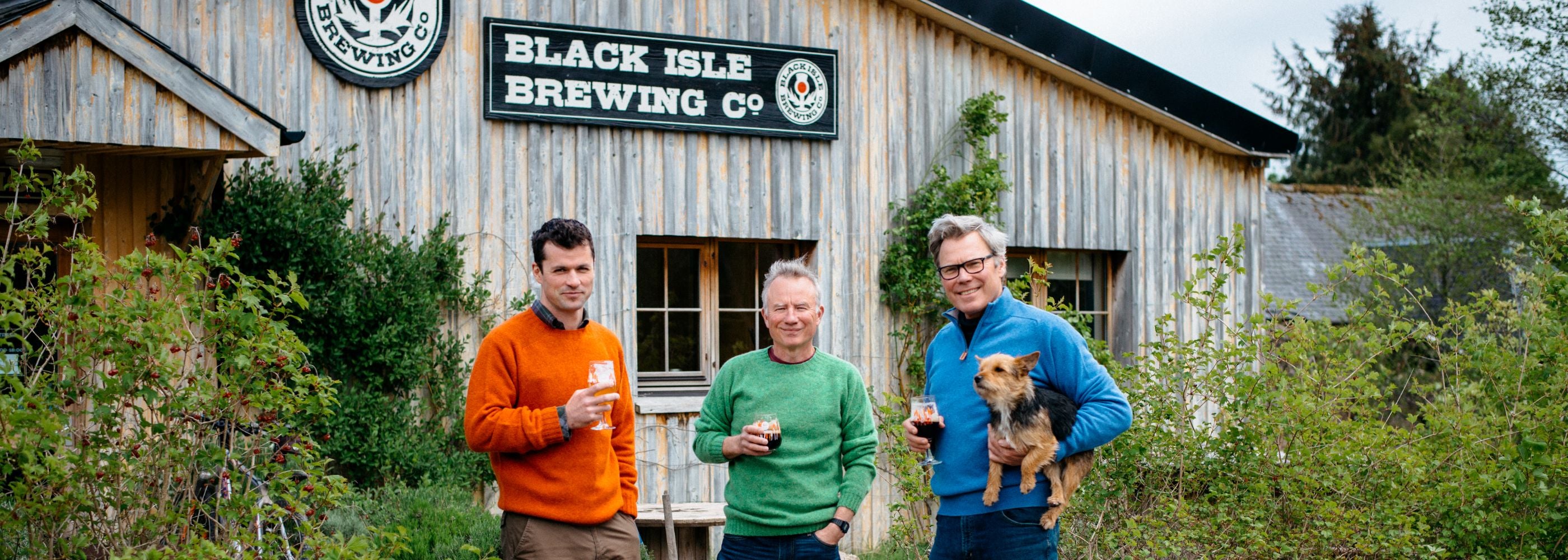 Three men stand outside Black Isle Brewing Co., each holding a drink. One man holds a small dog. They are dressed in colorful sweaters and stand in front of a rustic wooden building surrounded by greenery.