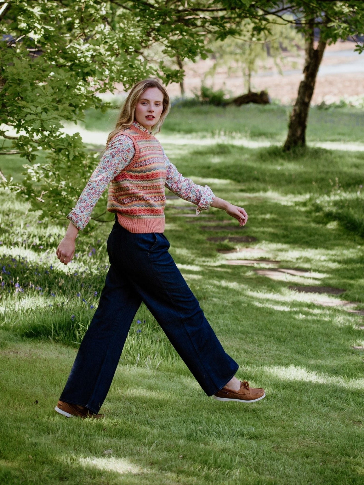 A person with long blonde hair walks on a grassy path in a sunlit park, wearing a patterned sweater vest over a floral shirt, brown shoes, and Campbell's of Beauly Auld Stock Wide Leg Jeans. Trees and leafy branches surround the scene.